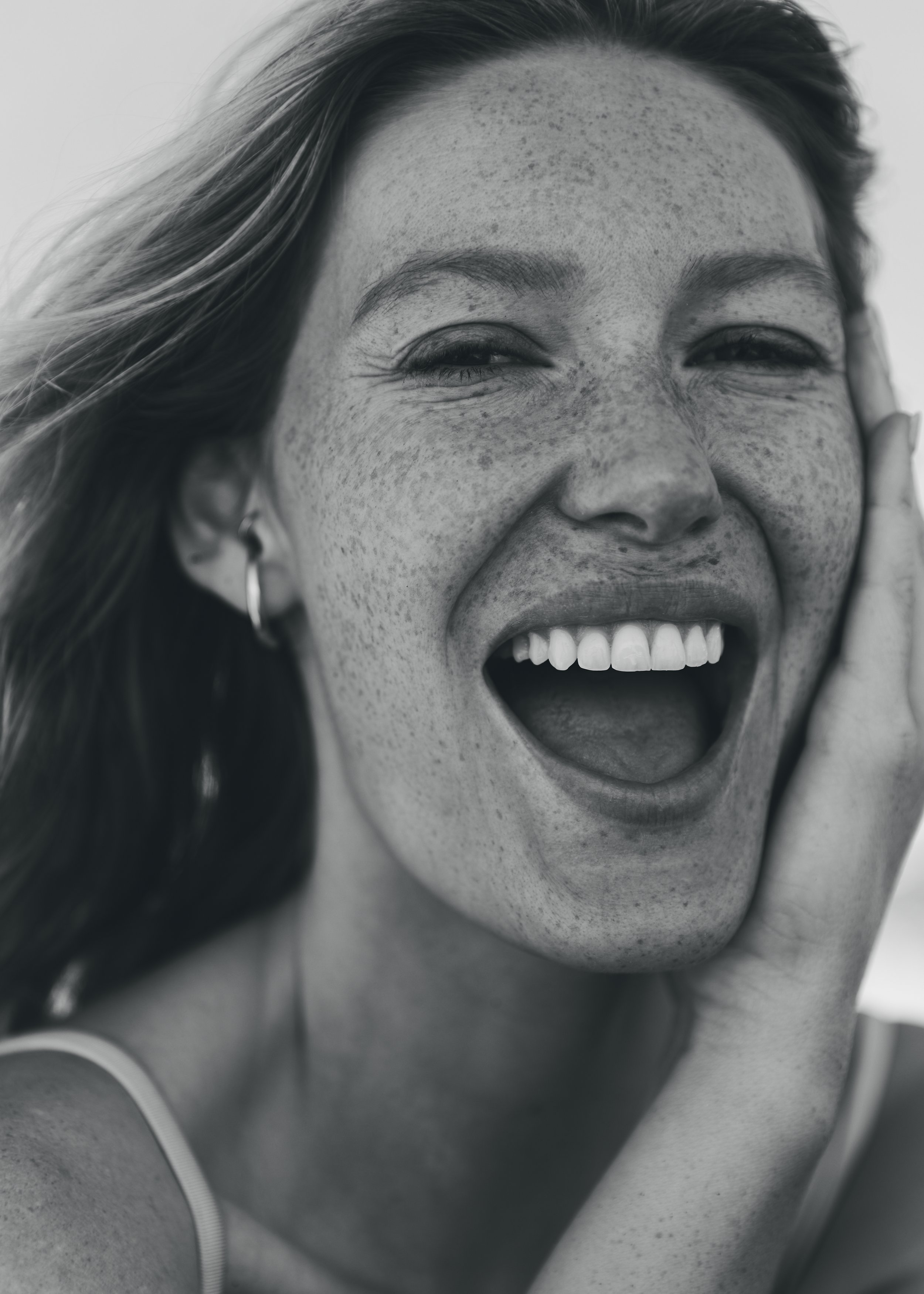 Close-up black and white photo of a smiling woman with freckles, long hair, and hoop earrings, showing her teeth and tongue out.