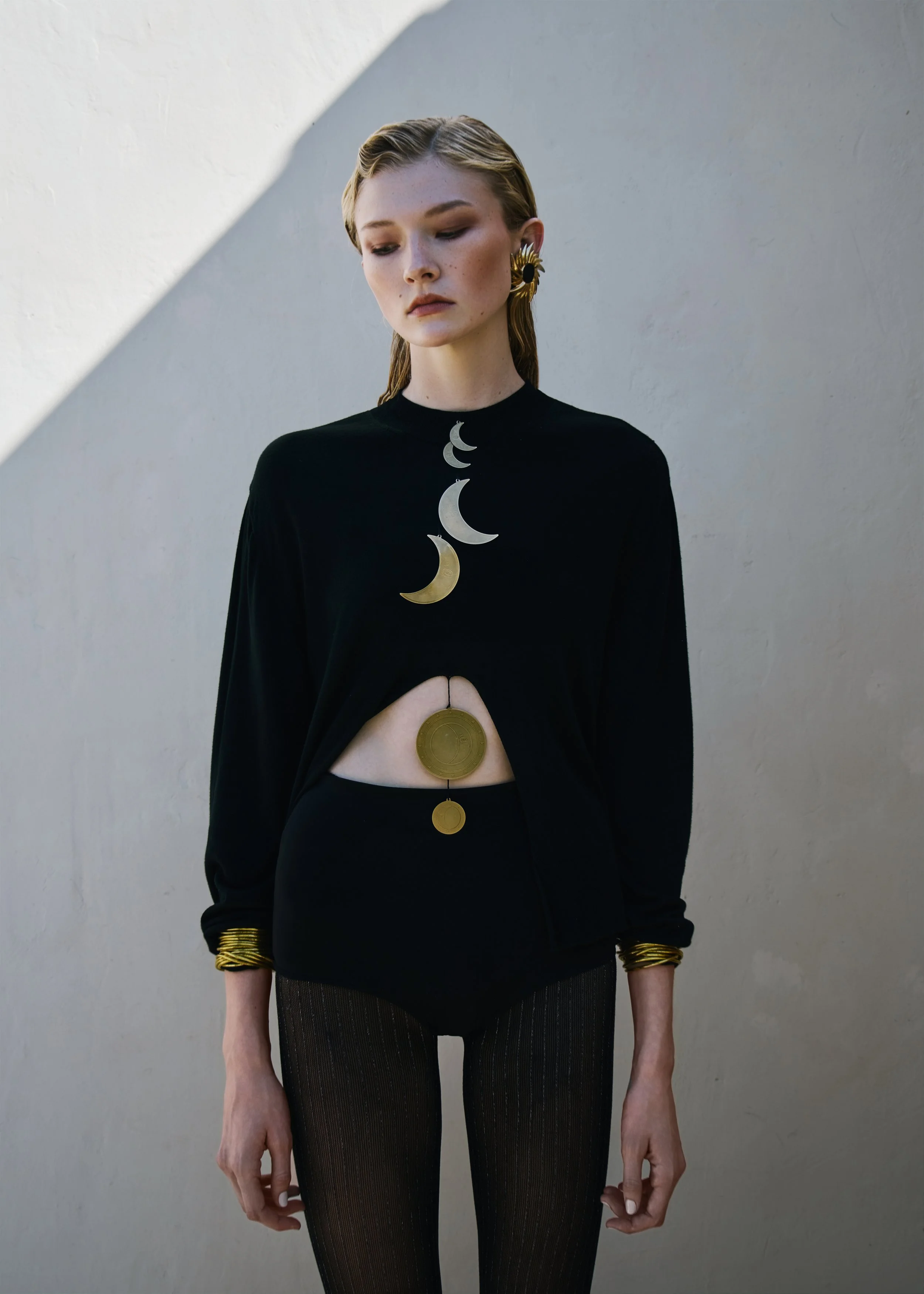 A woman with blonde hair and fair skin wears a black top decorated with a large moon phase necklace, gold earrings, and gold bangles, standing against a plain wall.