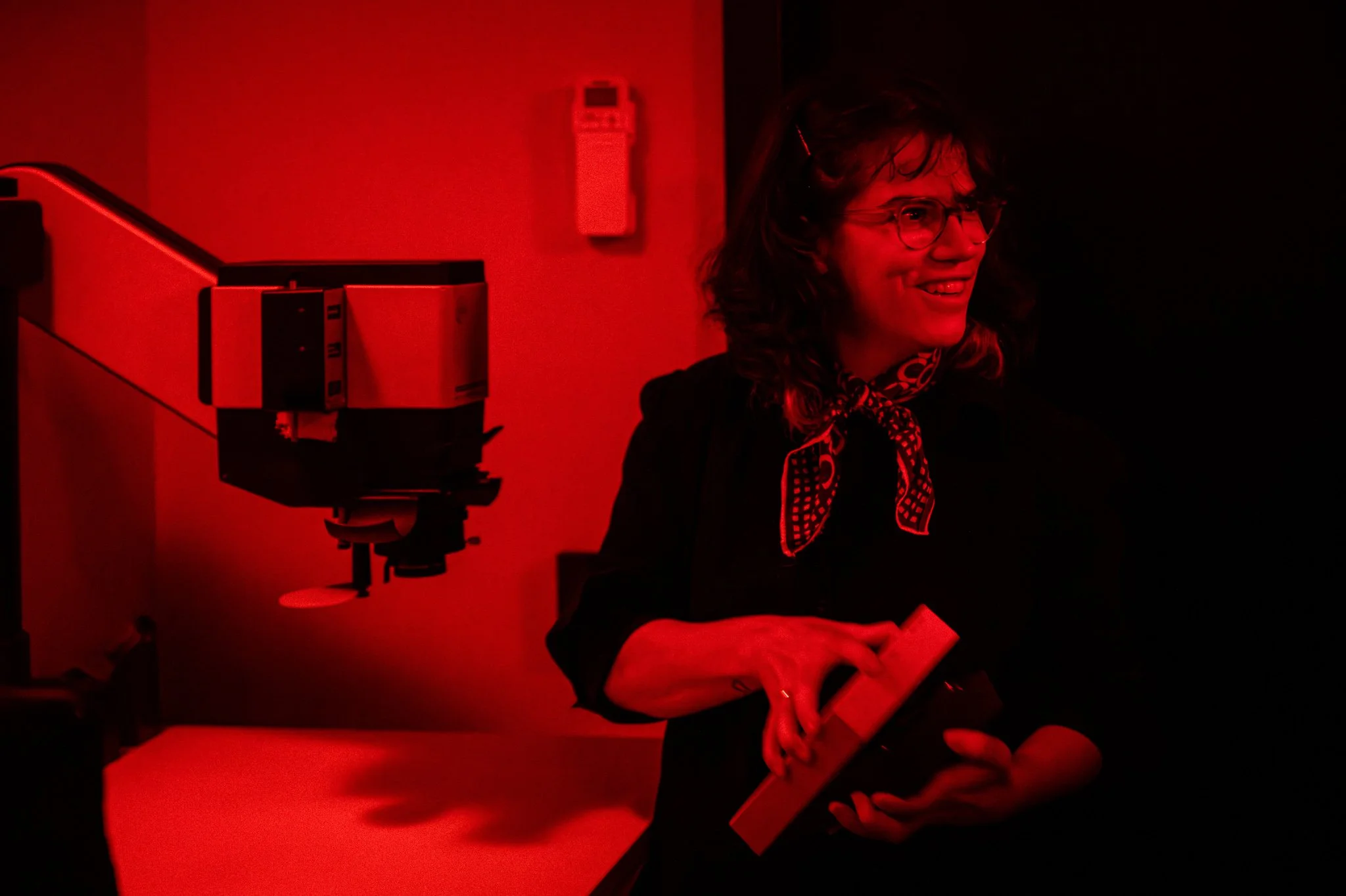 Analog photography darkroom showing a woman opening a box with light sensitive paper in a darkroom with an enlarger.