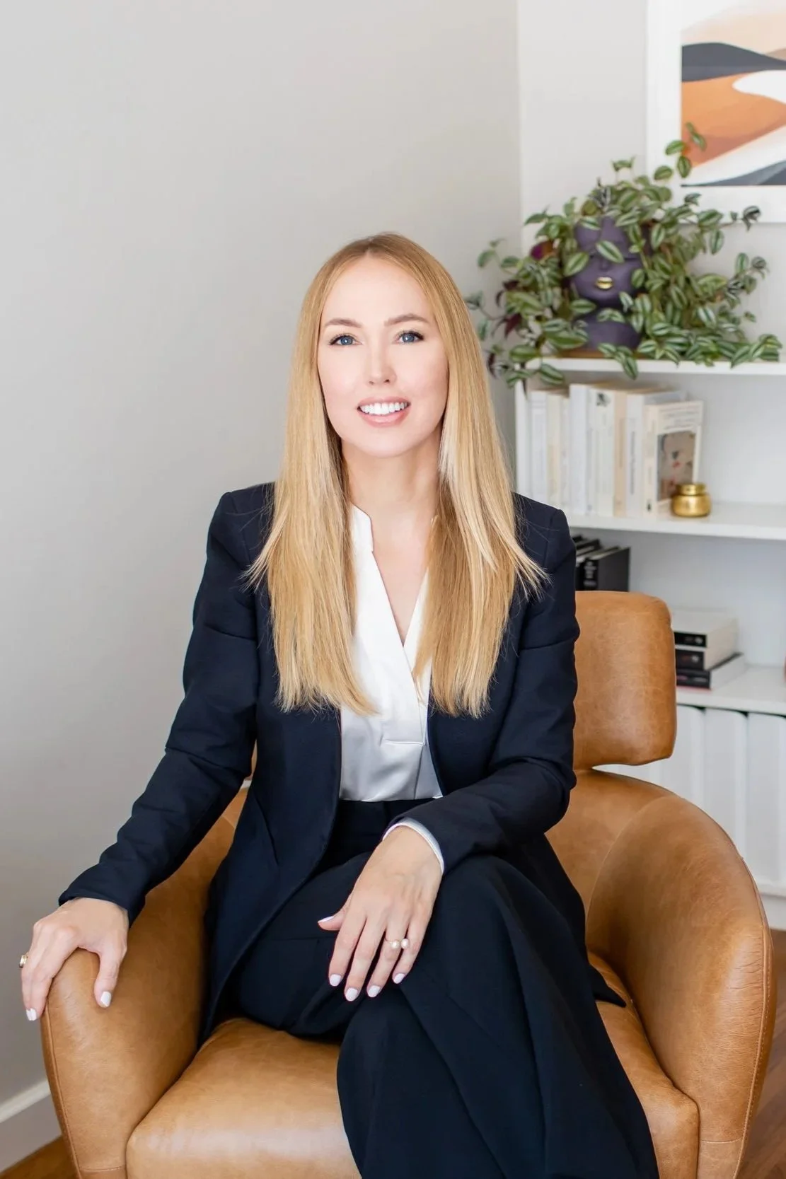 Dr. Sarah Stevens, PsyD sitting in a tan leather swivel chair with a bookcase, plant, and wall art behind her