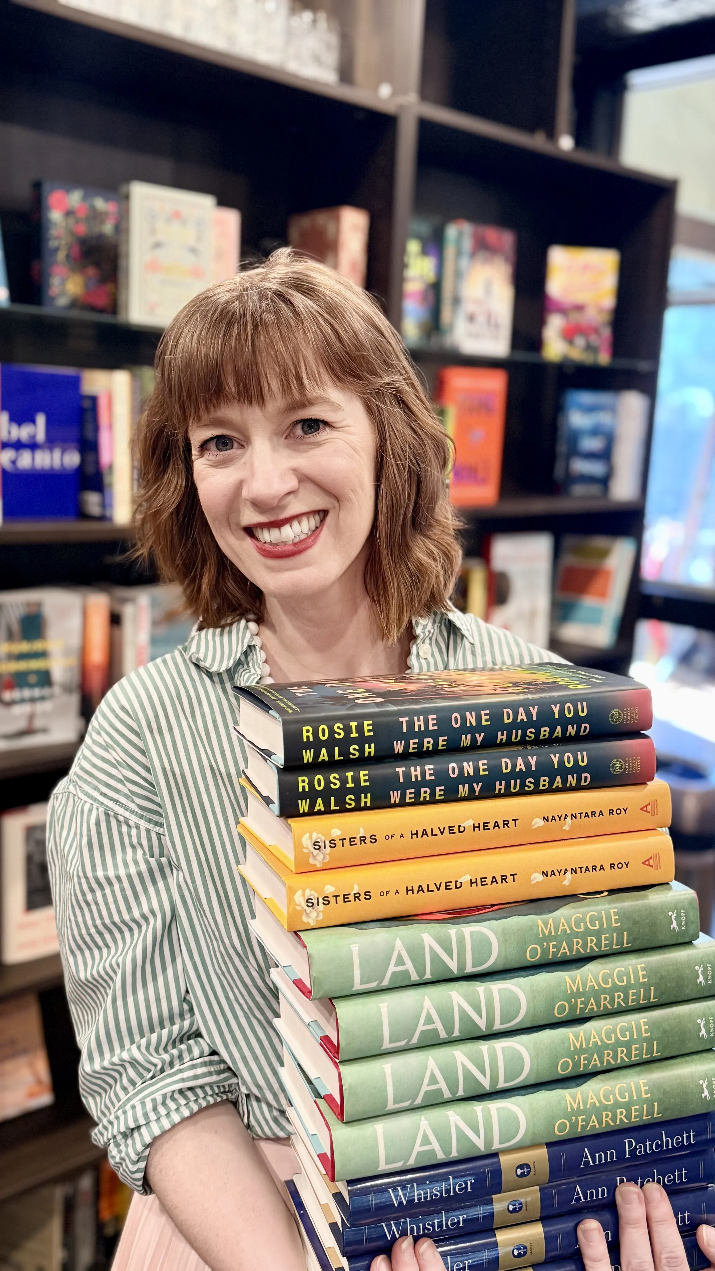 A smiling woman with shoulder-length reddish-brown hair and wearing a green and white striped shirt is holding a large stack of books in a bookstore.