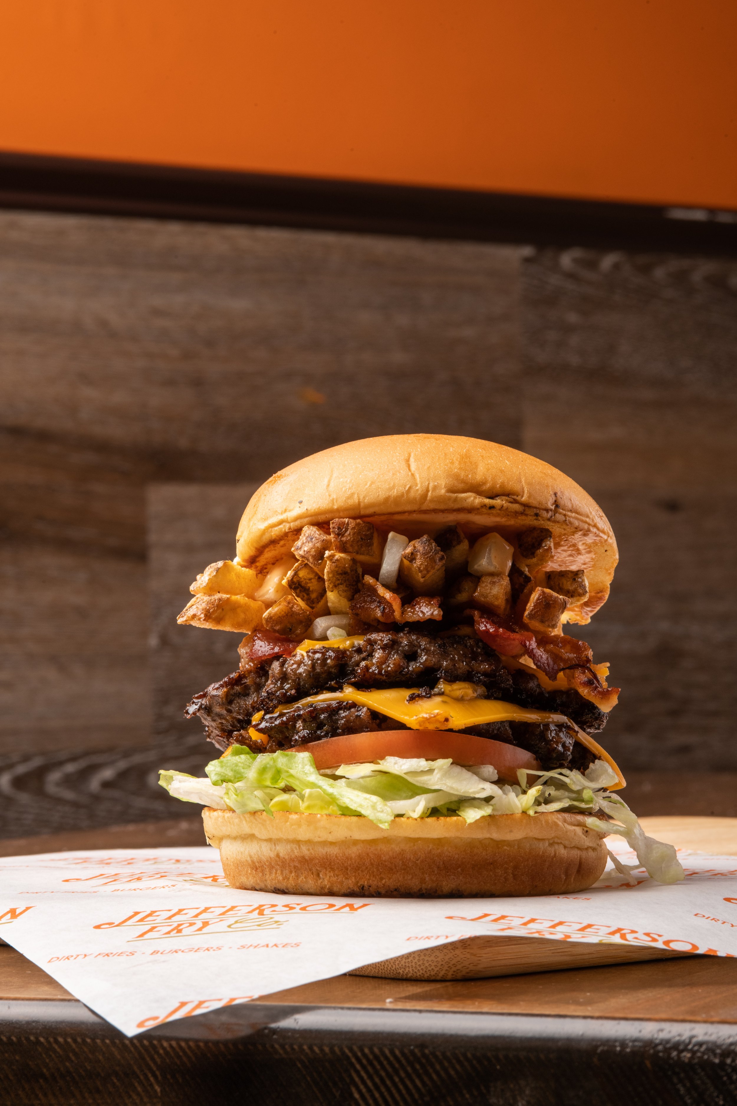 A cheeseburger with lettuce, tomato, cheddar cheese, two beef patties, bacon, diced onions, fried potatoes, and a bun, placed on a paper wrap on a wooden surface.