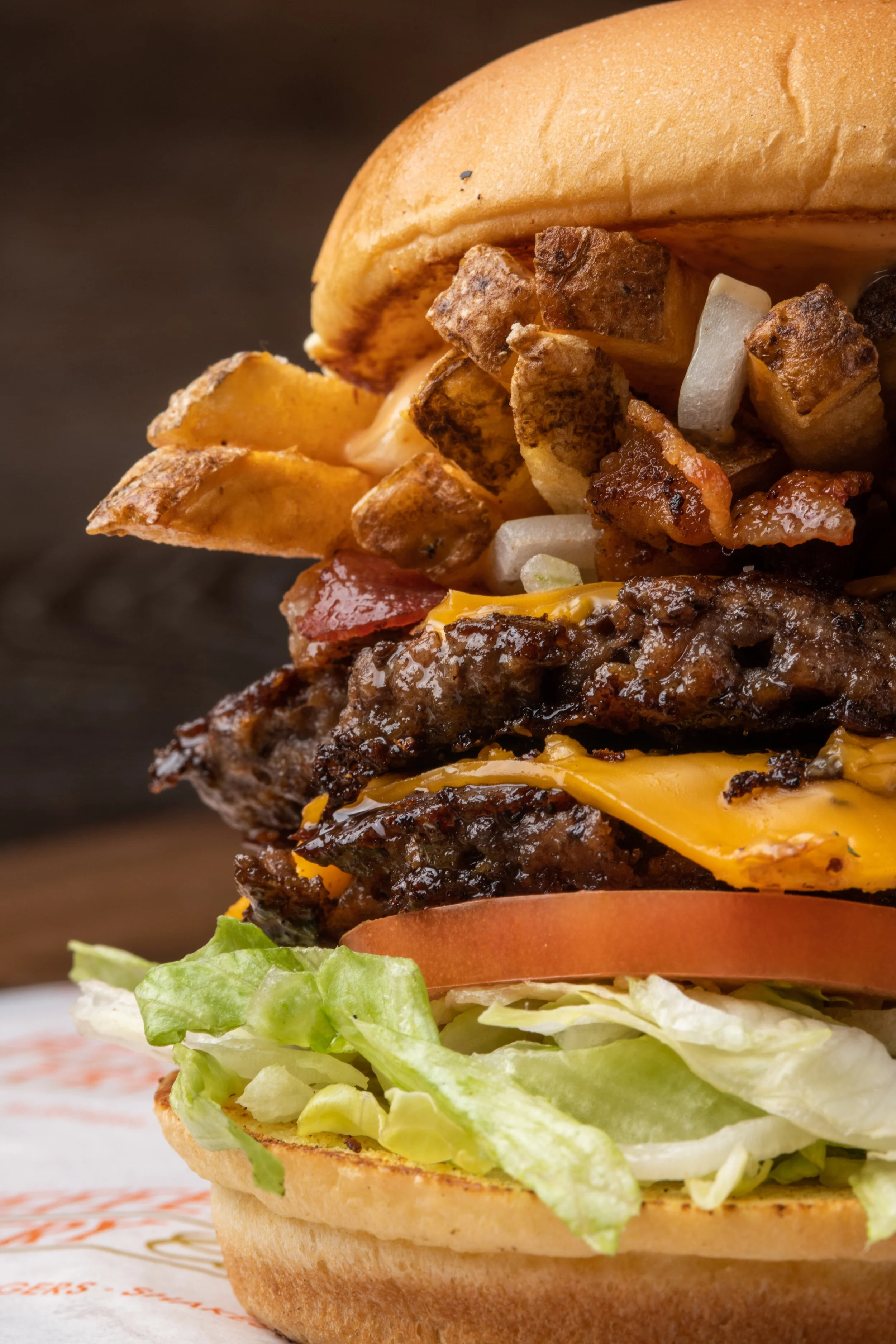 Close-up of a cheeseburger with lettuce, tomato, grilled beef patty, melted cheese, bacon, fried potato wedges, and a toasted bun.