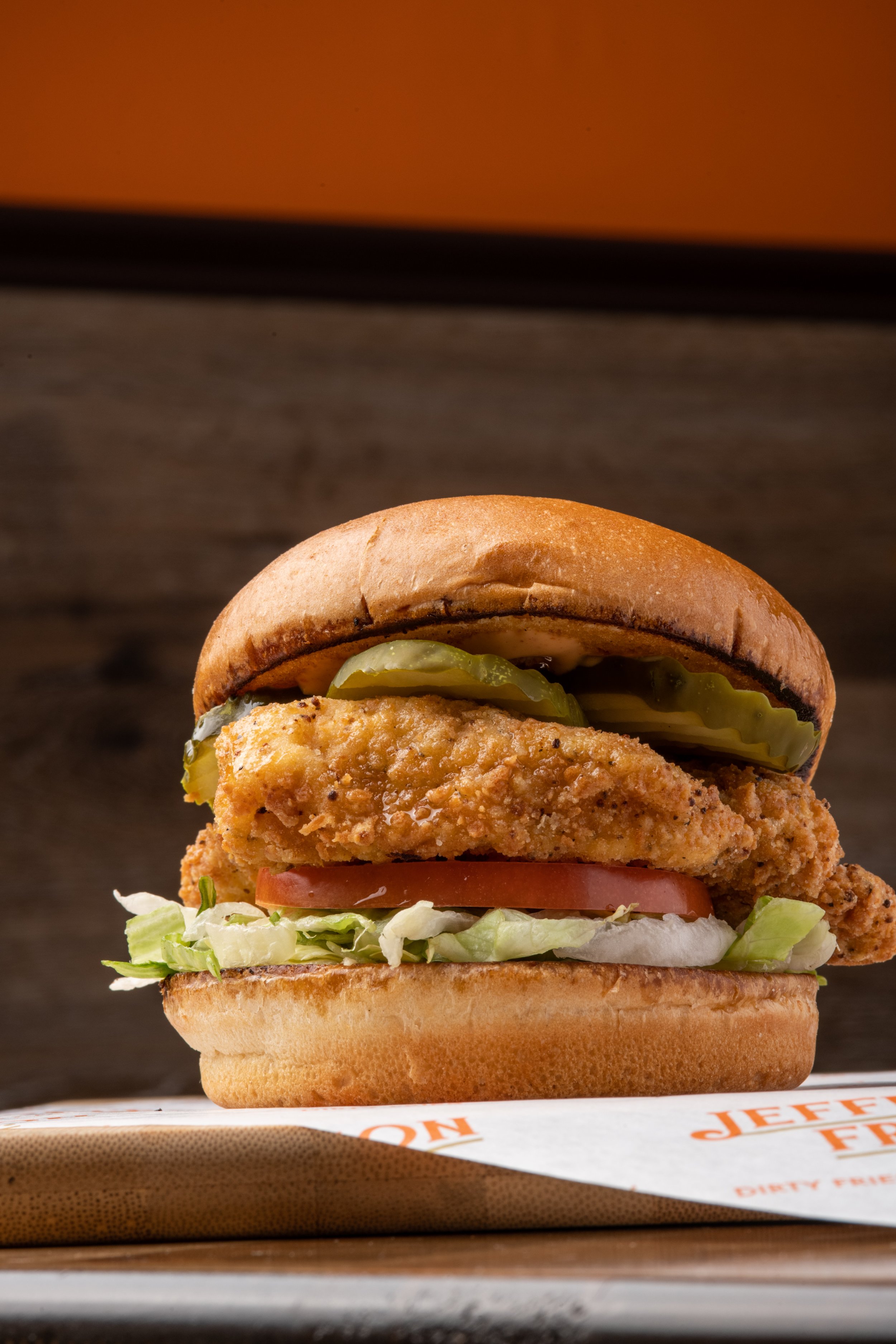 Close-up of a fried chicken sandwich with pickles, lettuce, tomato, and sandwich bun placed on a tray.