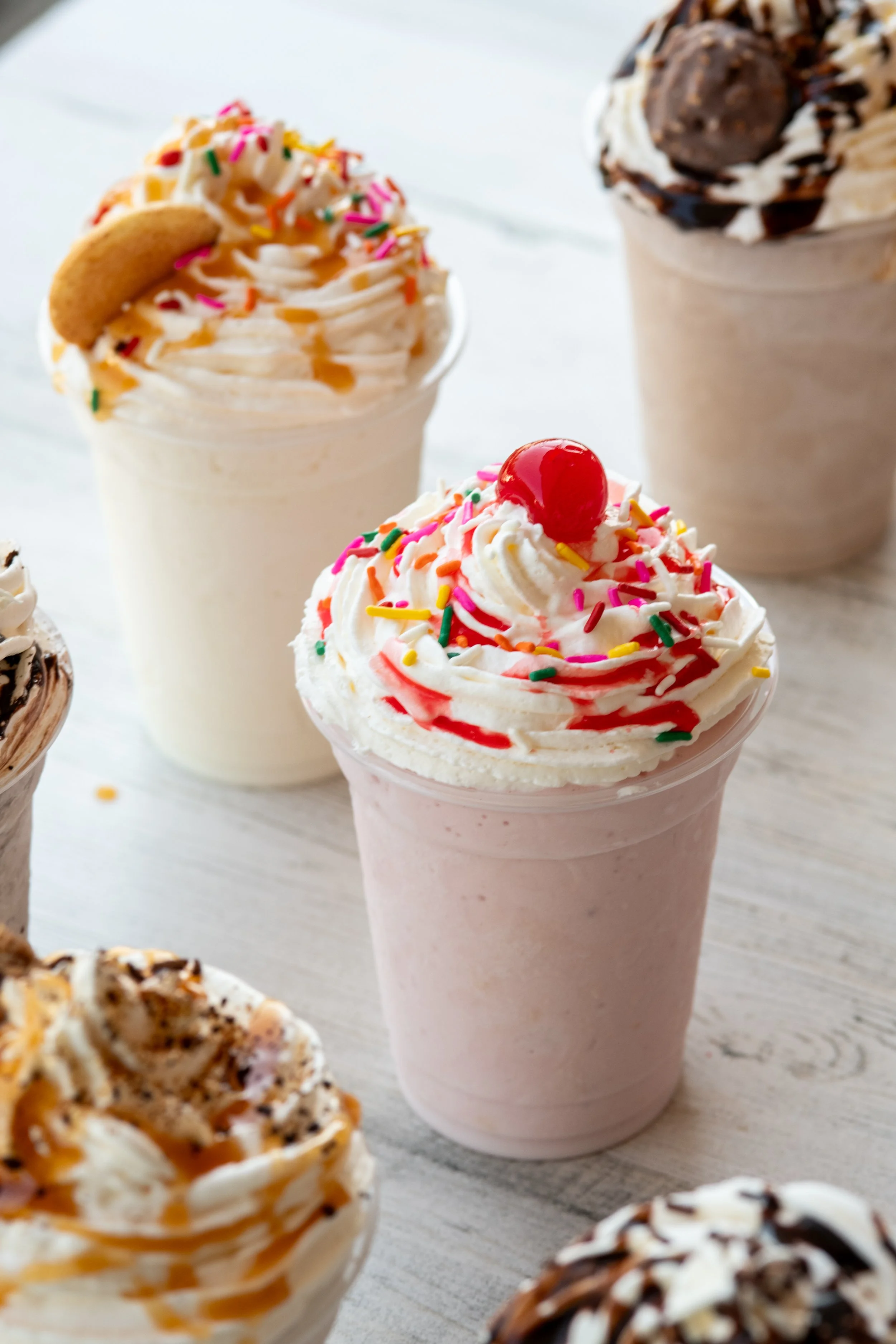 Four milkshakes with whipped cream, sprinkles, and toppings on a light gray wooden surface.