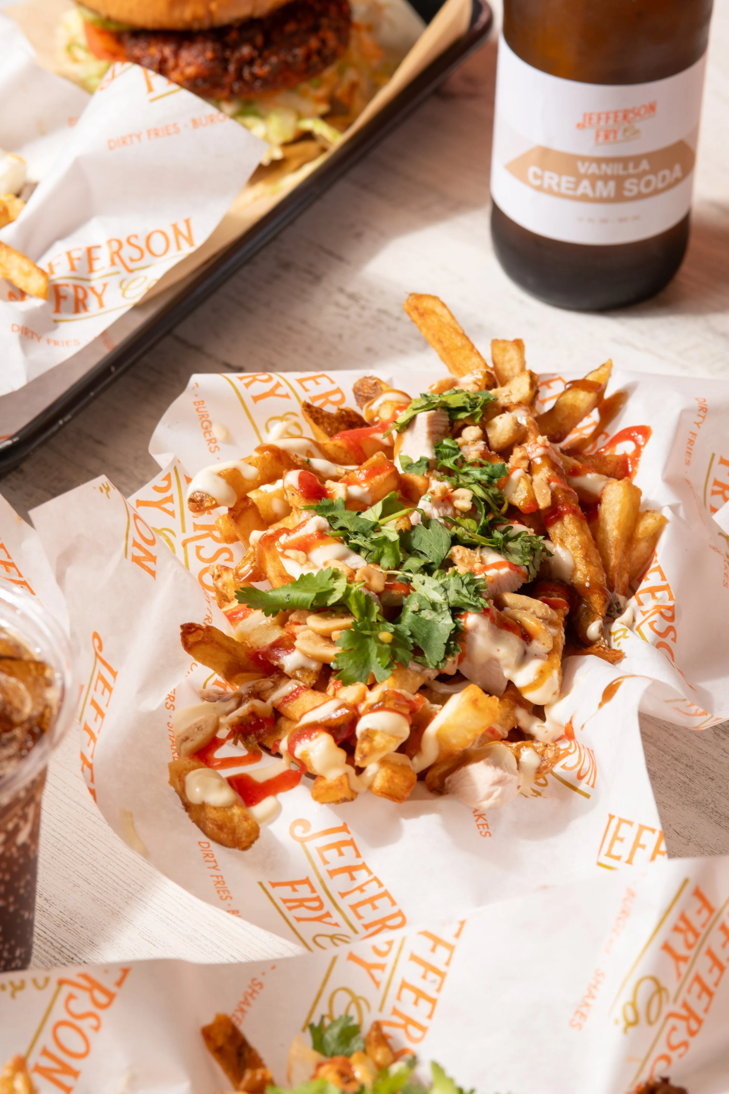 A serving of loaded fries topped with cilantro, chopped peanuts, and drizzled with white and red sauces on branded parchment paper from Jefferson Fry Co.