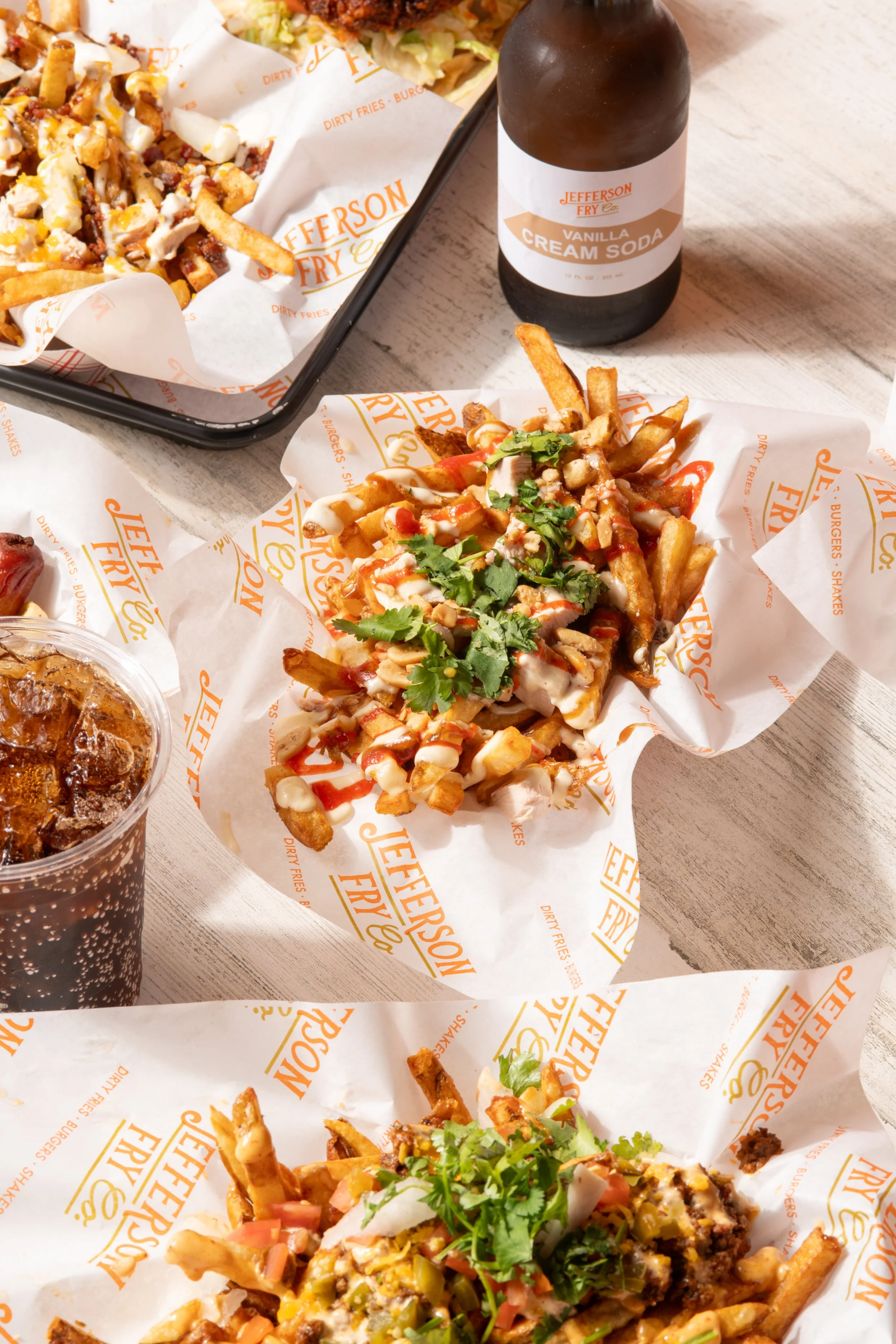 Several dishes of loaded fries with toppings like cheese, sauces, and herbs, a glass of soda, and a bottle of vanilla cream soda on a light-colored table.