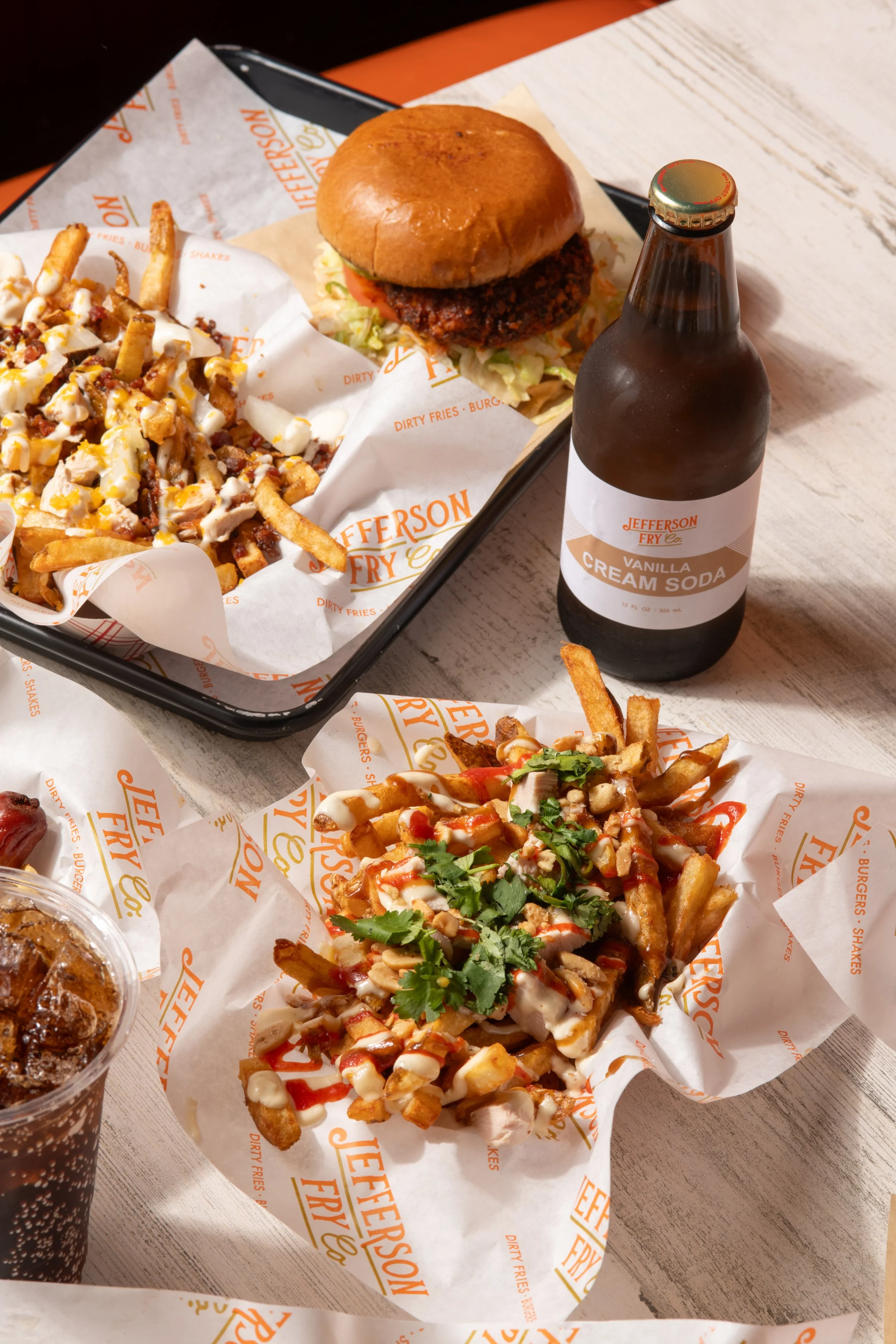 A tray with a burger, fries, and a vanilla cream soda, along with another serving of loaded fries topped with sauce and cilantro on parchment paper, on a white wooden table.