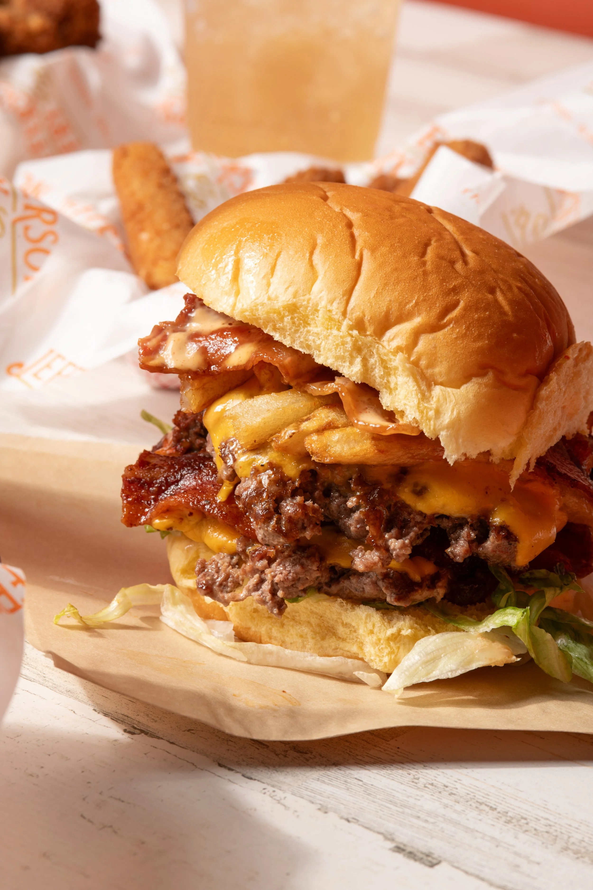 Close-up of a cheeseburger with lettuce, tomato, bacon, melted cheese, and grilled beef patty in a bun, with a side of fries and a beverage in the background.