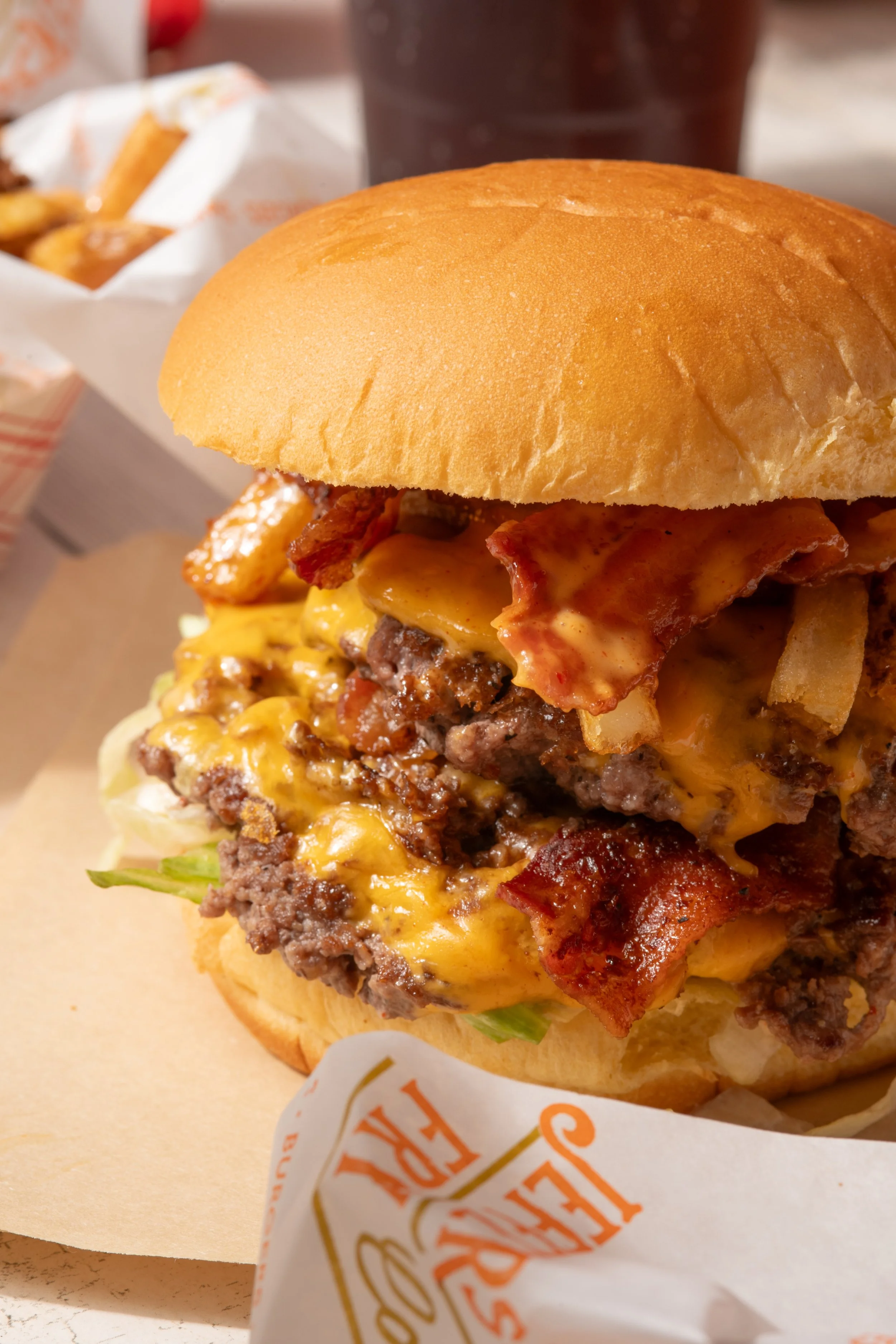 Close-up of a cheeseburger with bacon, lettuce, and melted cheese, served in a bun with a paper wrapper that says 'Etc. F'. In the background, there is a dark drink and a basket of fried chicken.