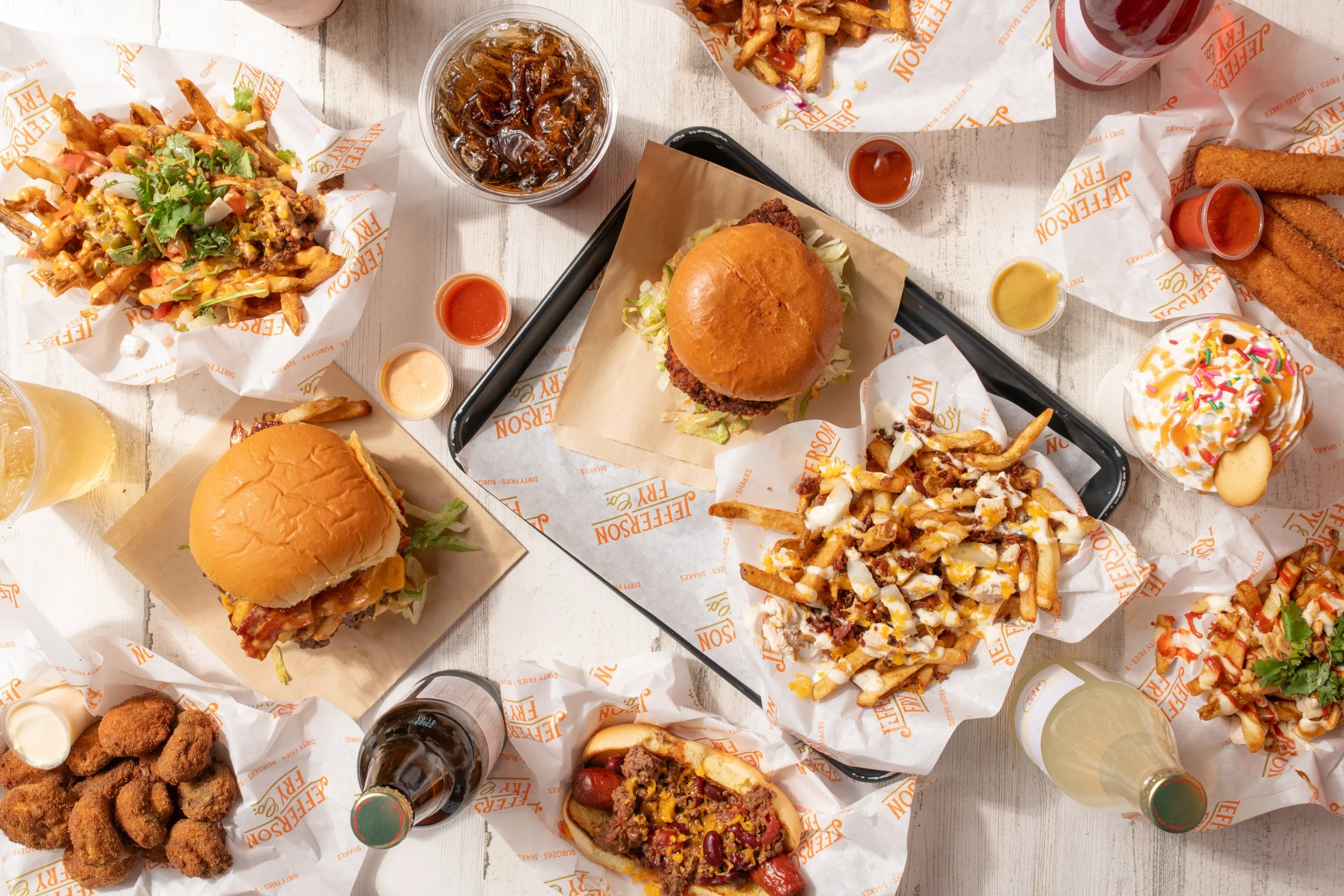 A table full of fast food including burgers, fries, fried chicken, onion rings, a hot dog, and various drinks and condiments.