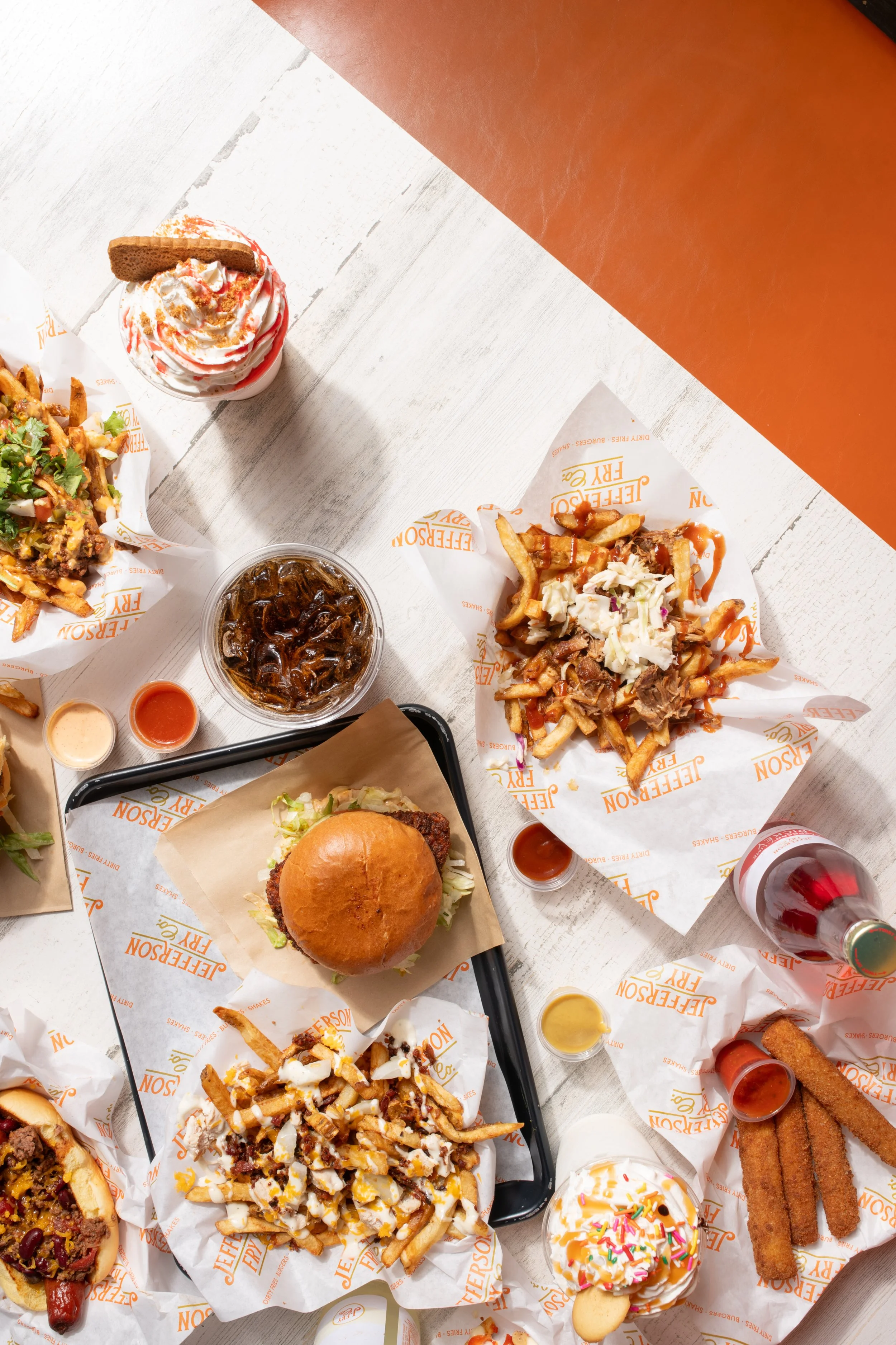 A top-down view of various fast food items on a white wooden table, including a burger, fries with toppings, a milkshake, soft drinks, chicken tenders, and desserts like ice cream and cake.