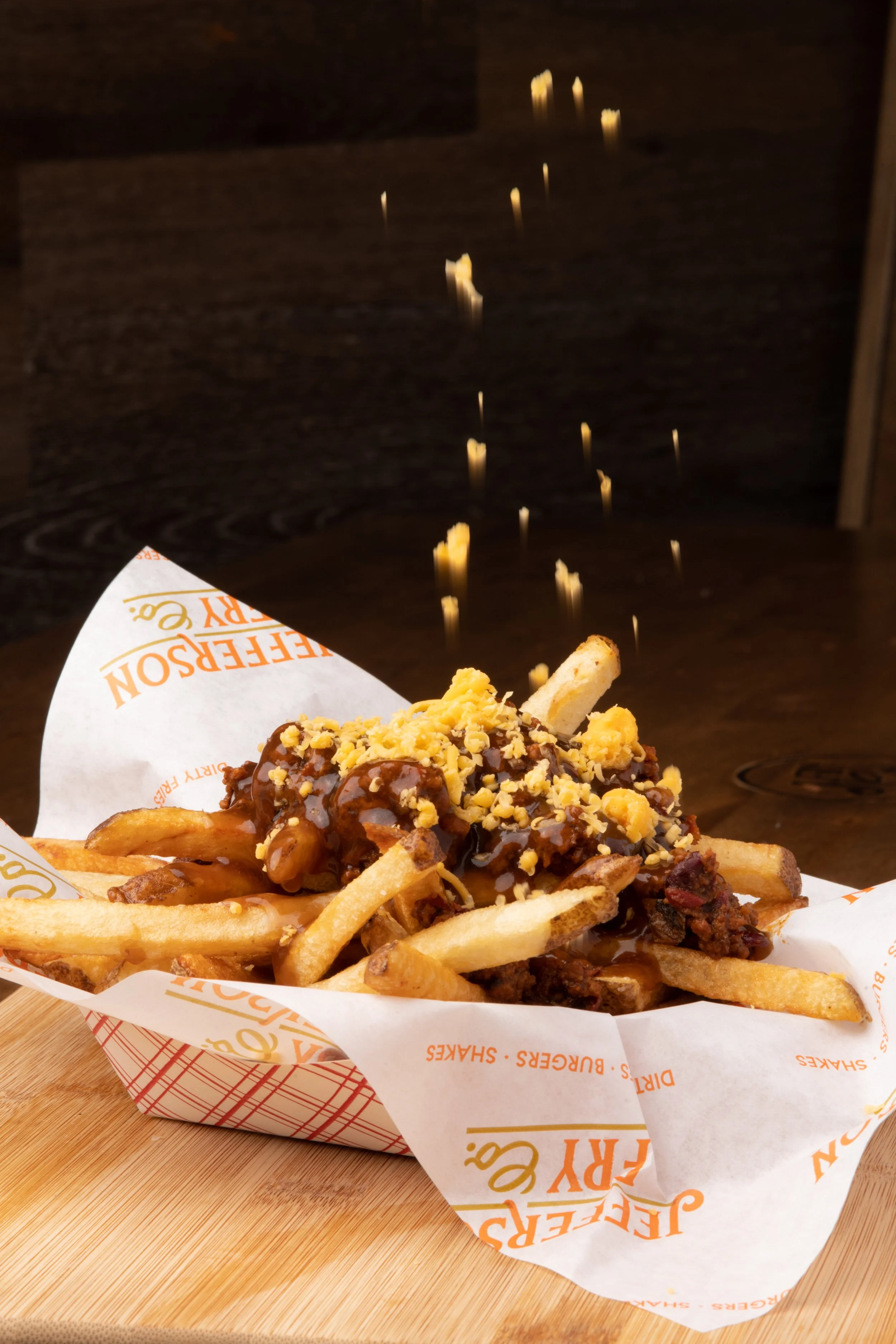A serving of chili cheese fries topped with shredded cheese, chopped nuts, and sauce, served in a paper boat on a wooden table with a dark background.
