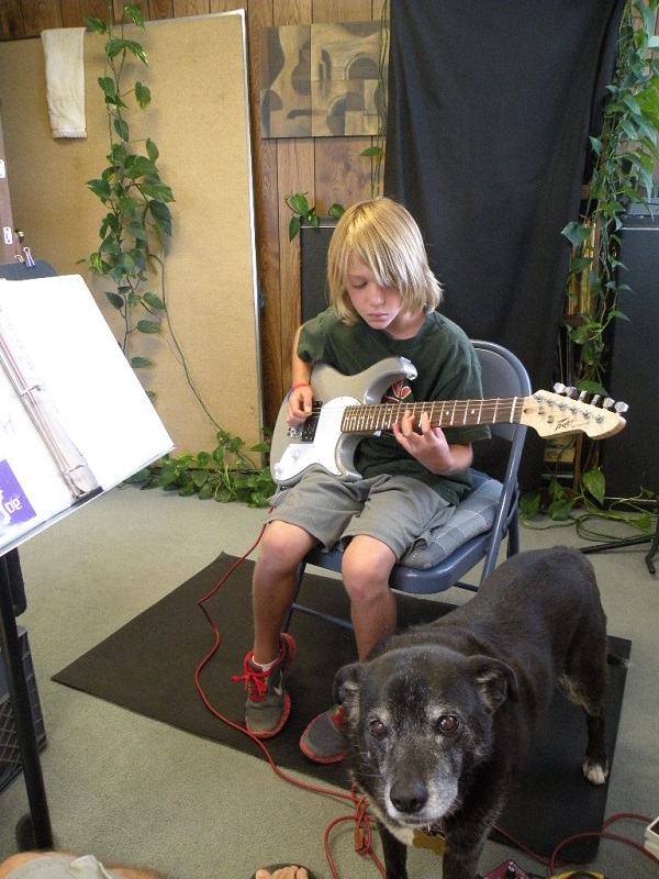    Young boy plays electric guitar while black mixed breed rescue dog stands by his side.   