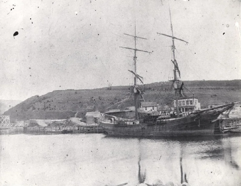 This vessel tied up at the harbour in Alma is probably being loaded with timber, the ship was used to transport the lumber to market. Note the wooden bridge across the Alma river in the far left of the photo, and the piles of logs ready to be sawed on the dock.