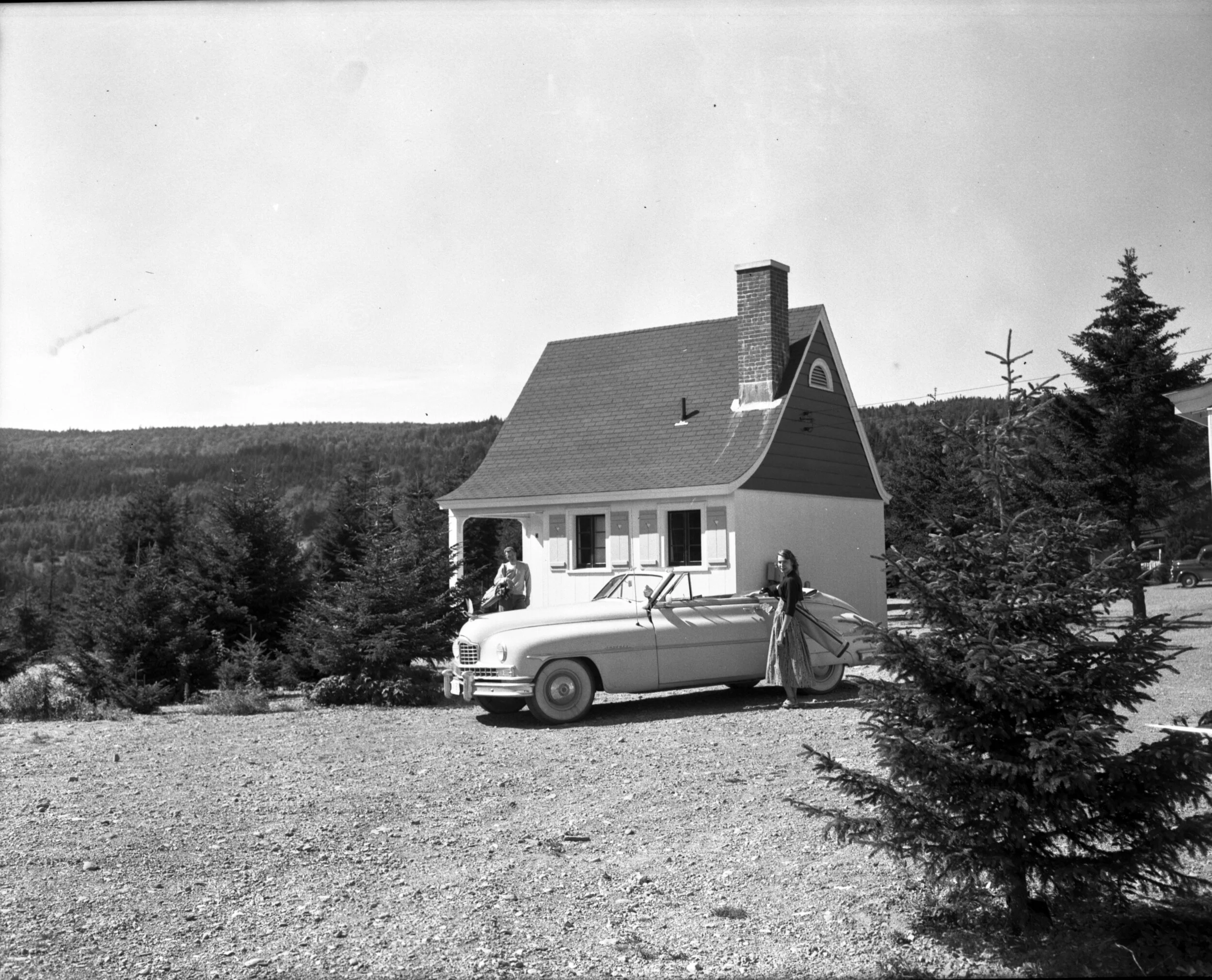 Couple leaving chalet with golf clubs over shoulder and convertible car parked in driveway, Fundy National Park, c.1950.From the New Brunswick Archives P93-AL-169
