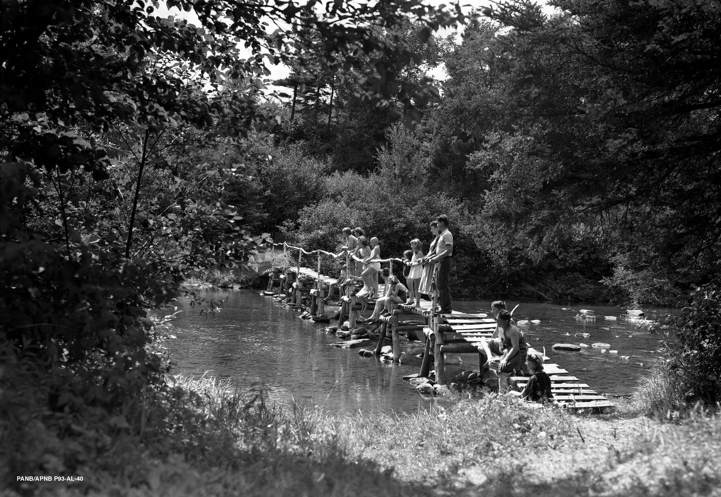 Group of people standing on wooden walkway crossing the river, Camp Hermit Thrush near Elgin, c.1940. From the New Brunswick Provincial Archives P93-AL-40