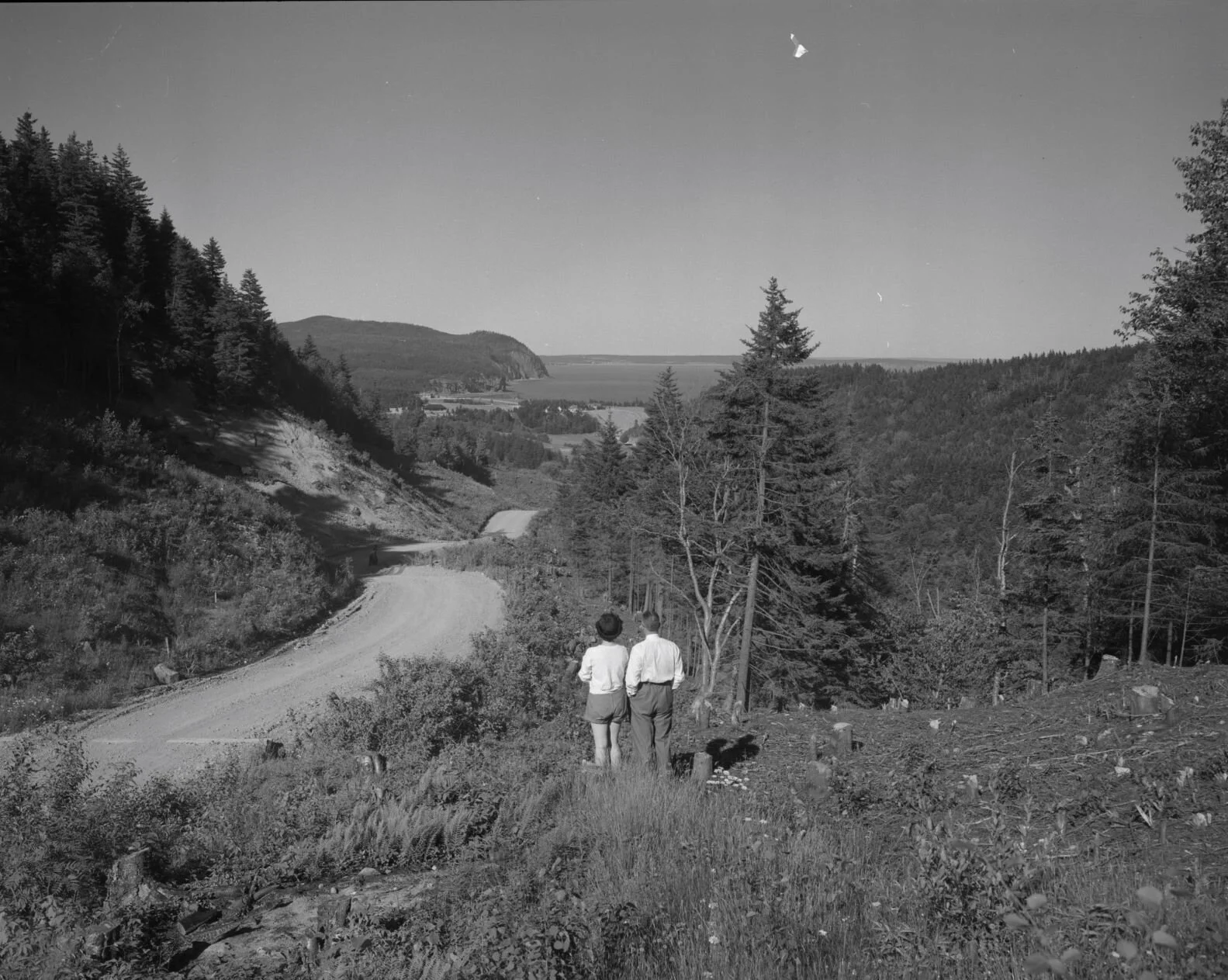 Two people standing looking down gravel road into valley of trees and waterfront, road approaching Fundy National Park, c. 1950.  From Archives New Brunswick P93-AL284..