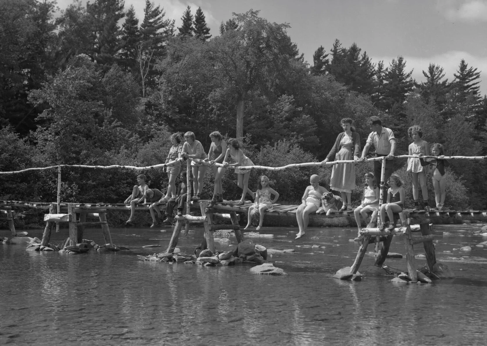 Group of people standing on wooden walkway crossing the river, Camp Hermit Thrush  near Elgin, c.1940.. From Archives New Brunswick. P93-AL-206