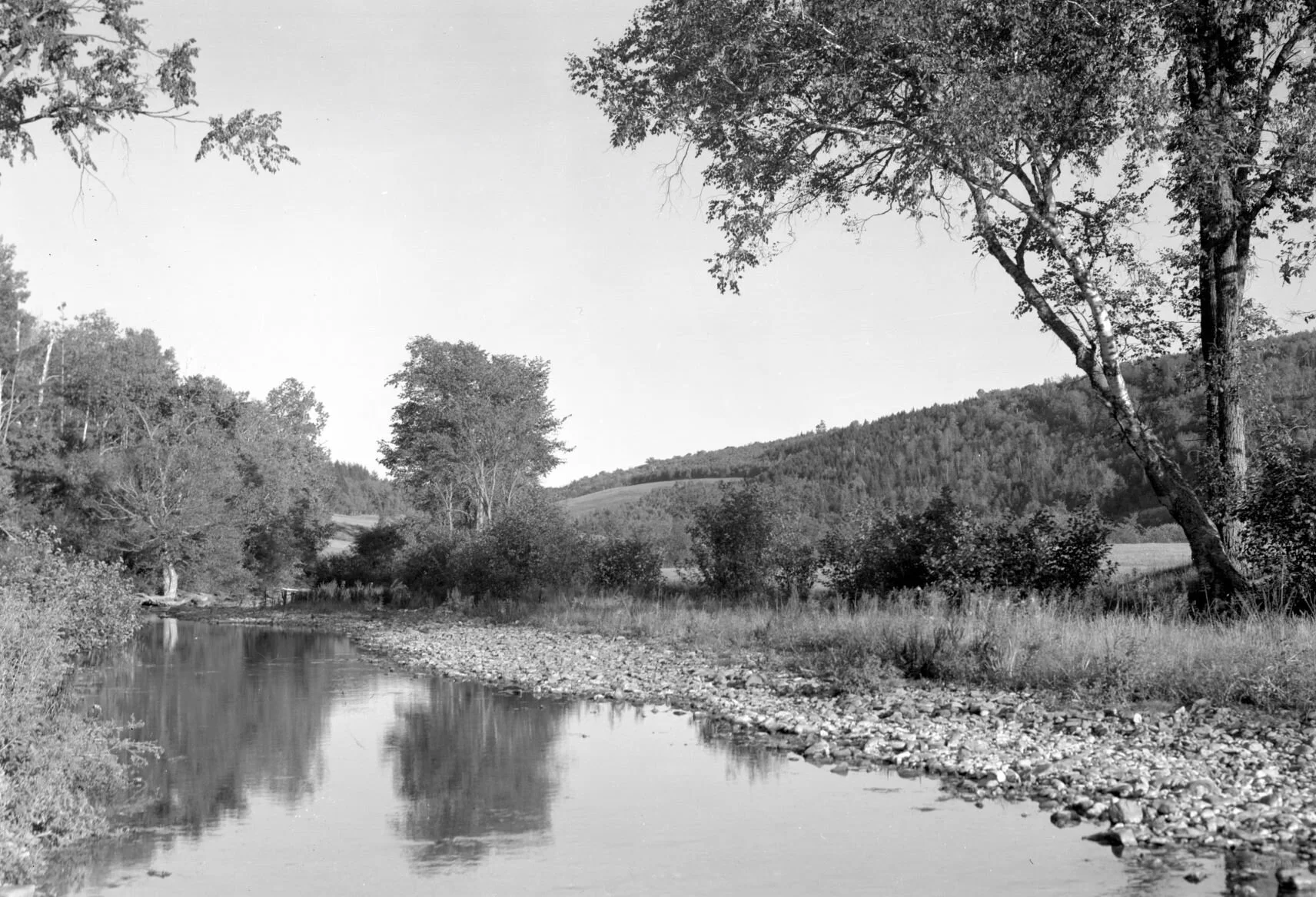 Shoreline of stream with hills and trees in background, Portage Vale, 1938. From Archives New Brunswick P93-AL-11