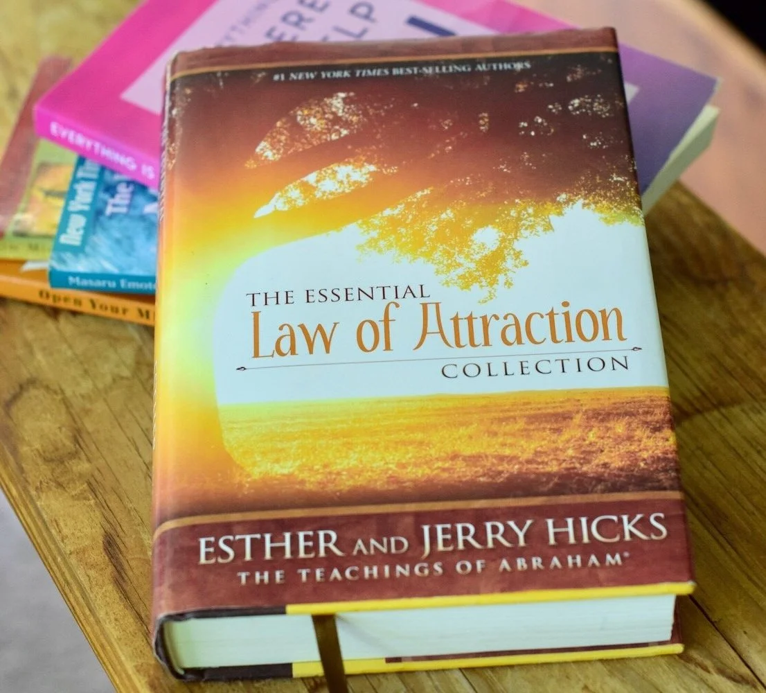 Photo of The Essential Law of Attraction Collection book, propped up on stack on books, Open Your Mind To Receive, The Four Agreements, and The Hidden Messages in Water on wooden table with chest in background by Amanda Linette Meder