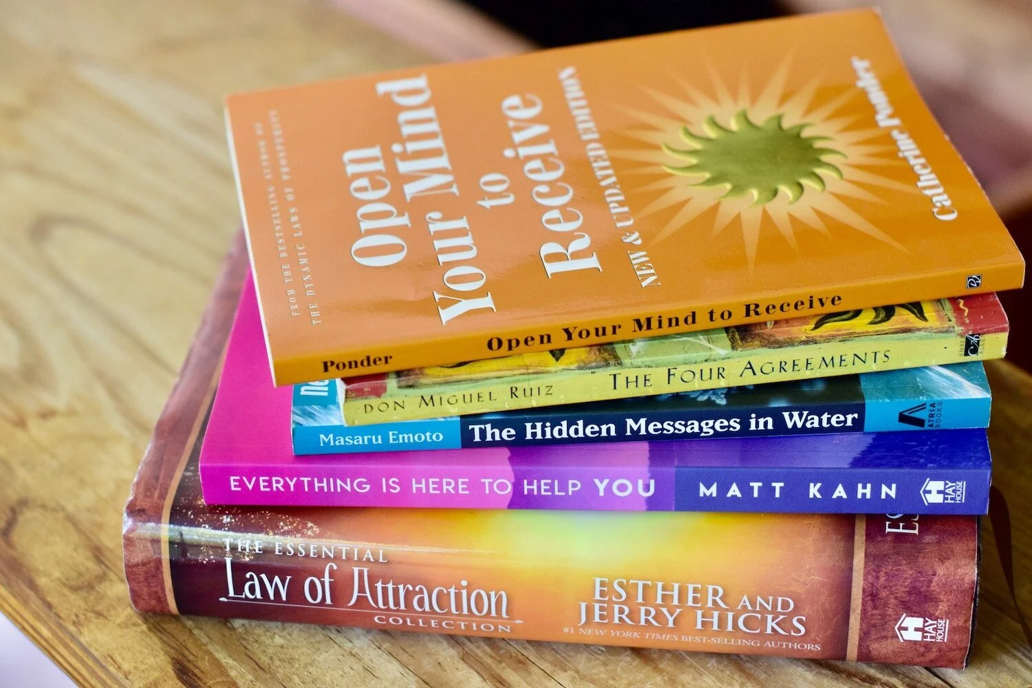 Photo of stack of books, Open Your Mind to Receive, The Four Agreements, The Hidden Messages in Water, Everything Is Here To Help You, The Essential Law of Attraction Collection on wooden table by Amanda Linette Meder