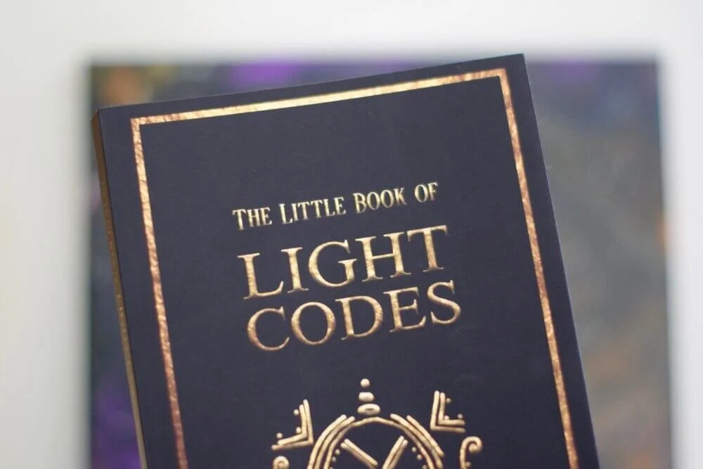 Gifted by Author - This image is a cover of The Little Book of Light Codes by Laara up against a galactic print by Amanda Linette Meder
