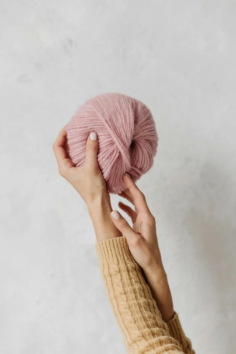 Hands holding a large ball of pink yarn in front of a plain wall.