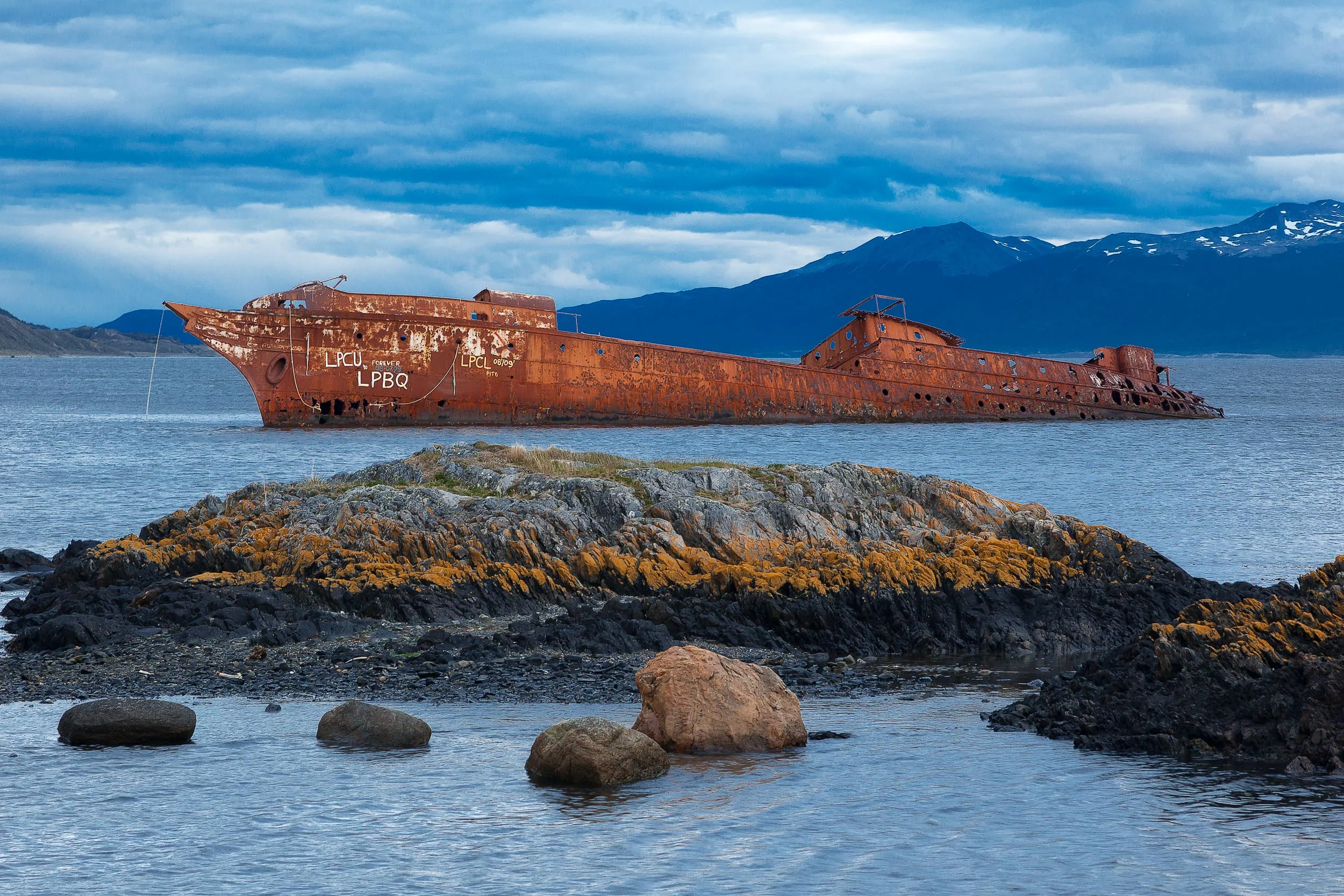 A ship, now no more than a rusting hulk , lies in a bay near the city of Ushuaia in the far south of Argentina . The orange color of the ship is illuminated by the gentle sunlight and is a striking contrast against the predominantly bluish light resulting from gathering storm clouds .