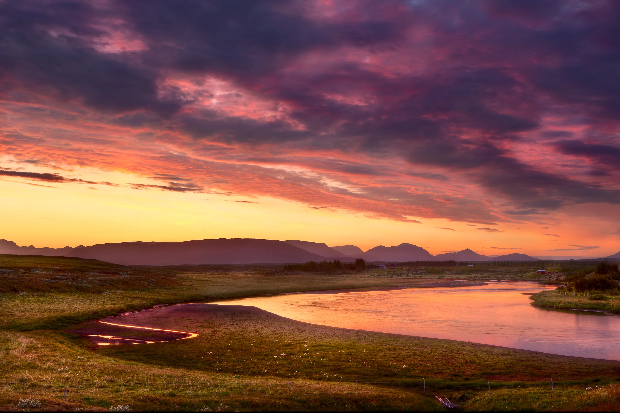 Sunset and a stormy sky reflect light onto a creek and farmland in rural Iceland .