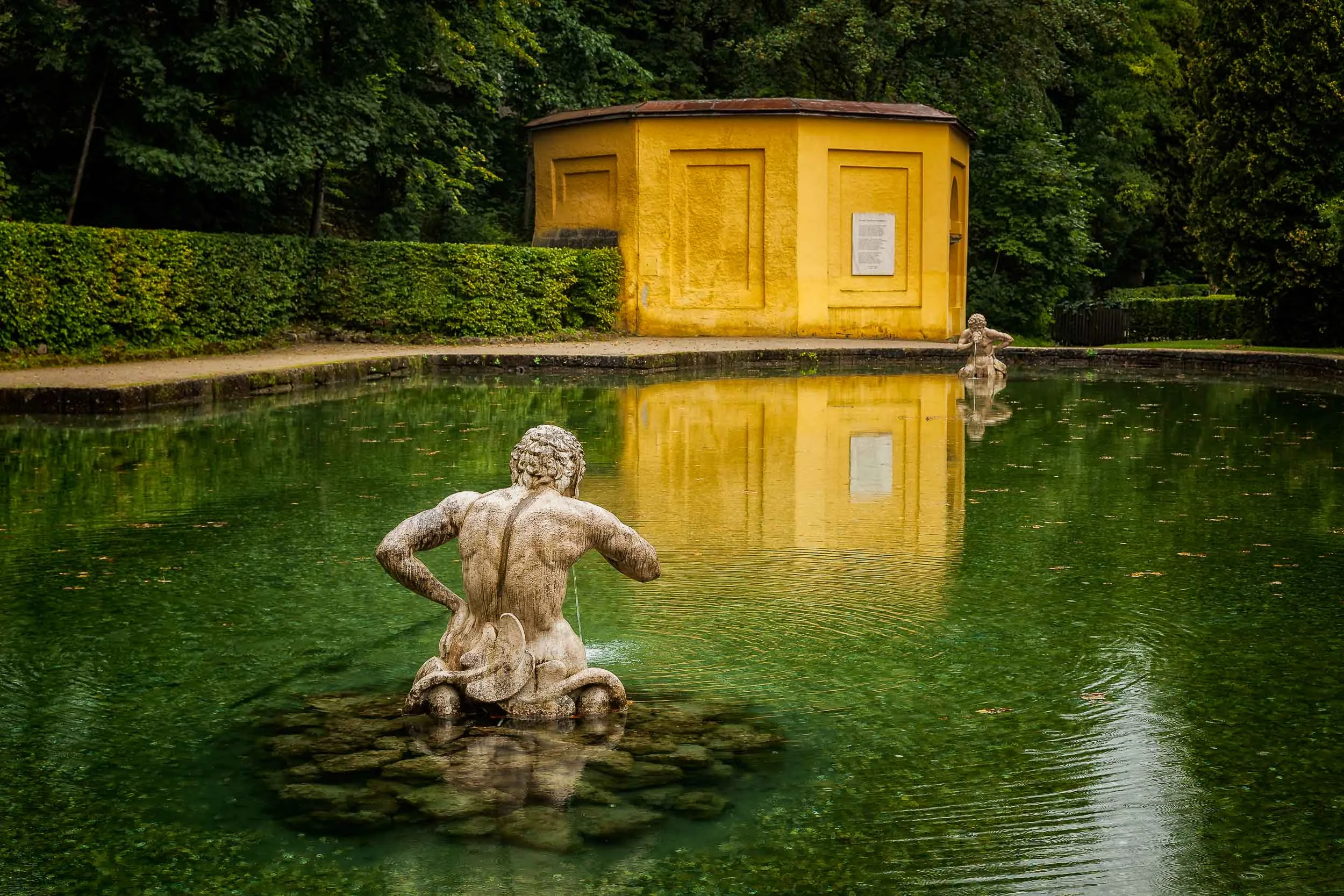 A bright yellow building and its reflection , between two statues in a pond, produce a serene scene in a lovely park setting in Salzburg, Austria .