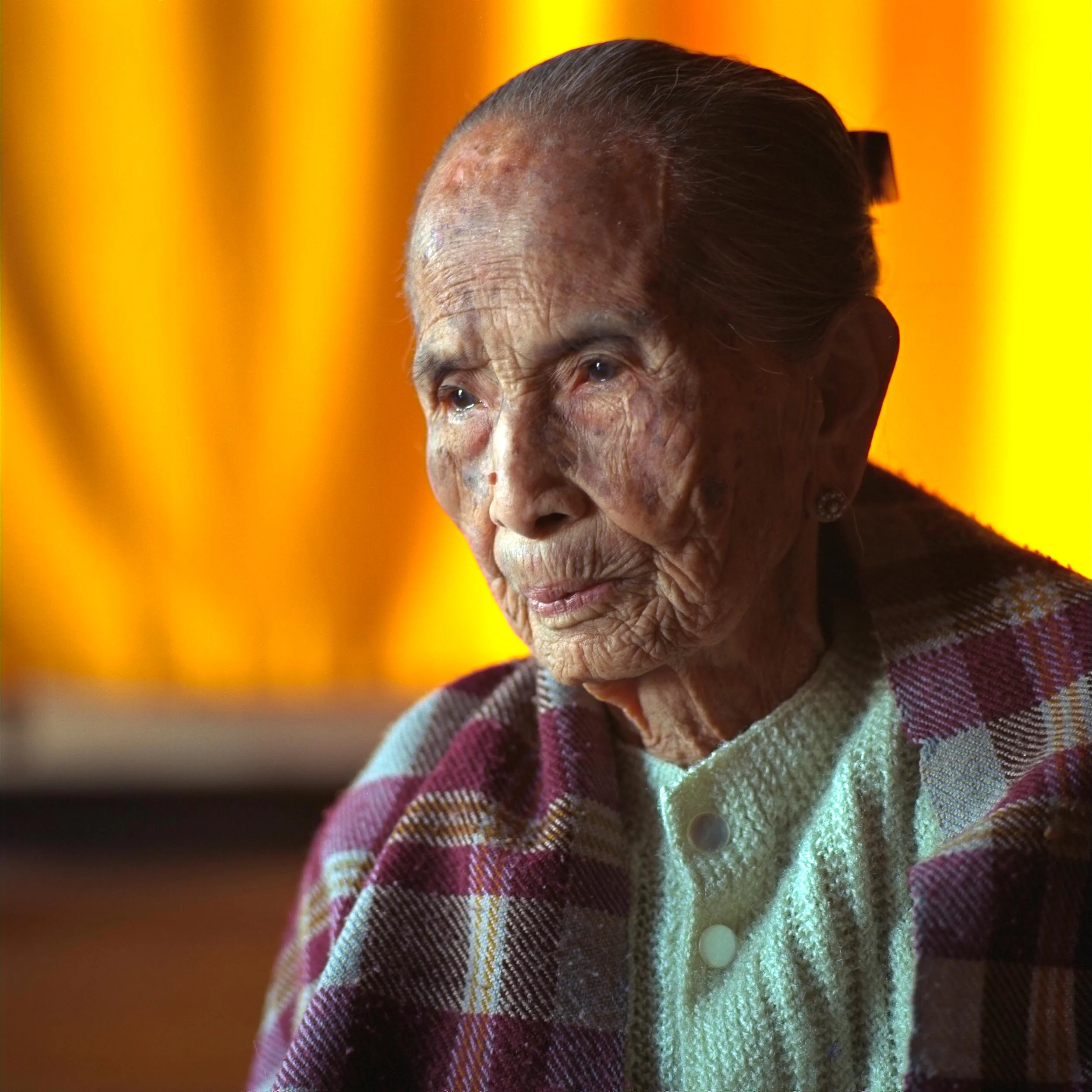 Portrait of an elderly woman against colorful curtains in her home in rural Myanmar .