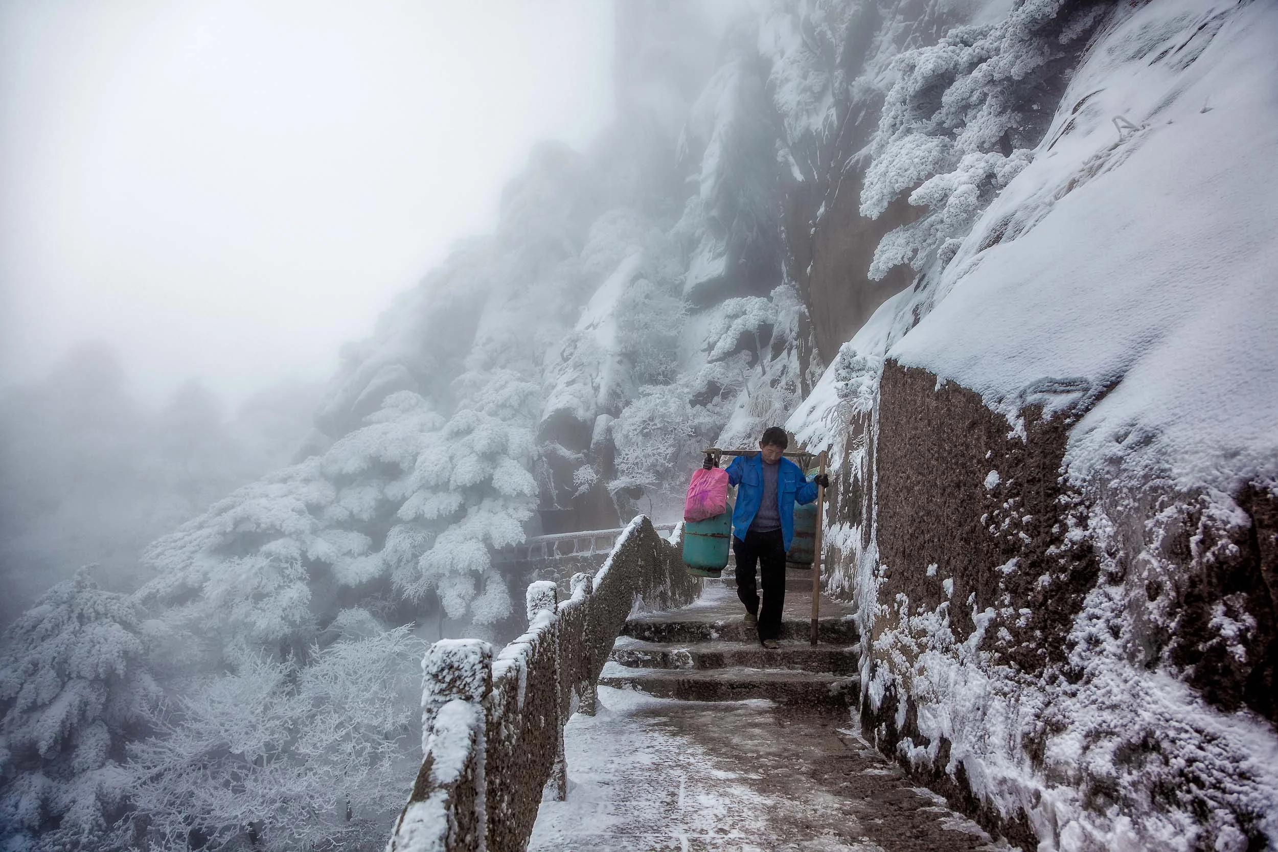 A porter , loaded with goods, walks a windy and exposed path on Huangshan (i.e., Yellow Mountain) in China .