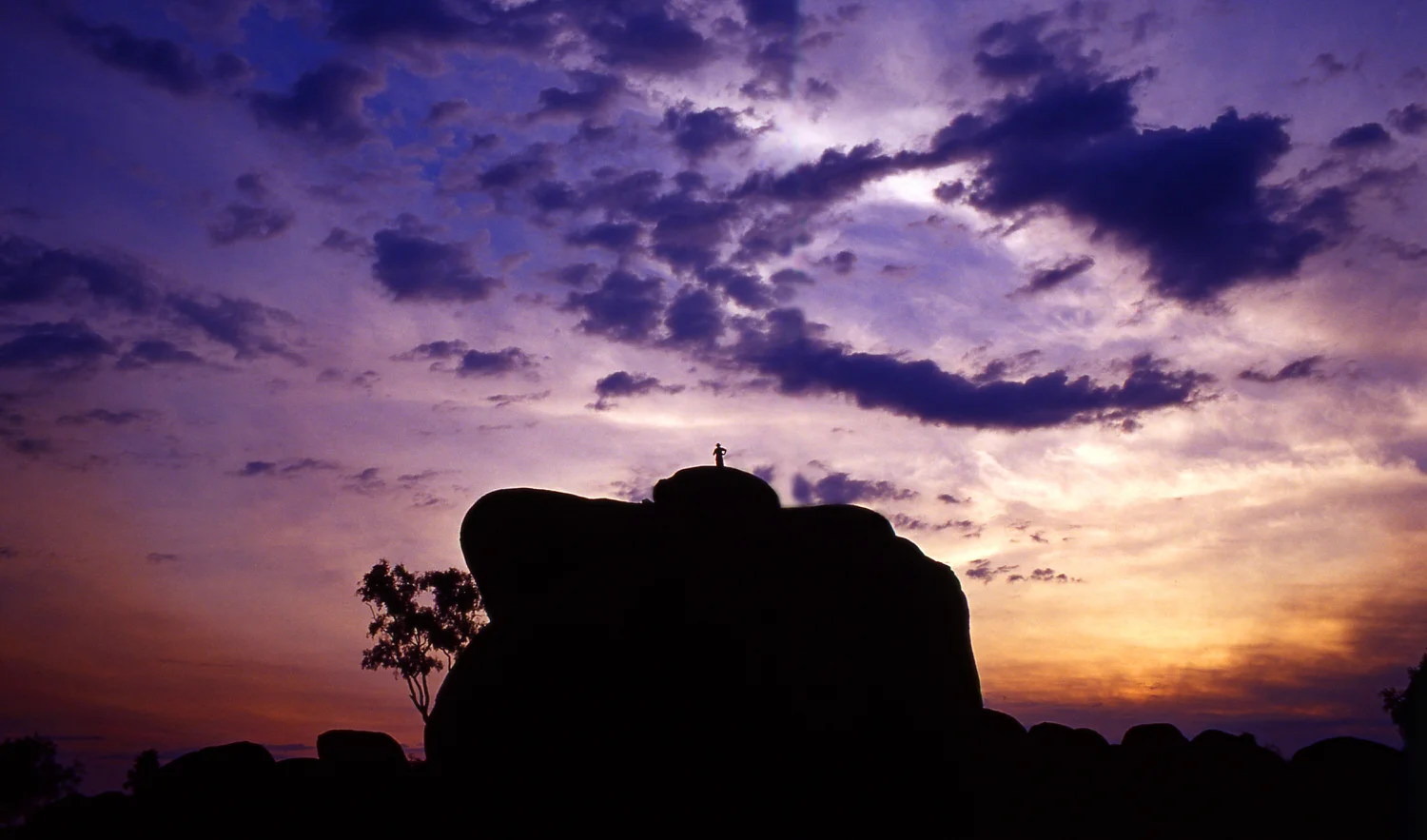 A tiny human figure, on top of a huge bolder backlit by a wonderous sunset, at Devils Marbles near the town of Wycliffe Well in the Northern Territory , Australia.