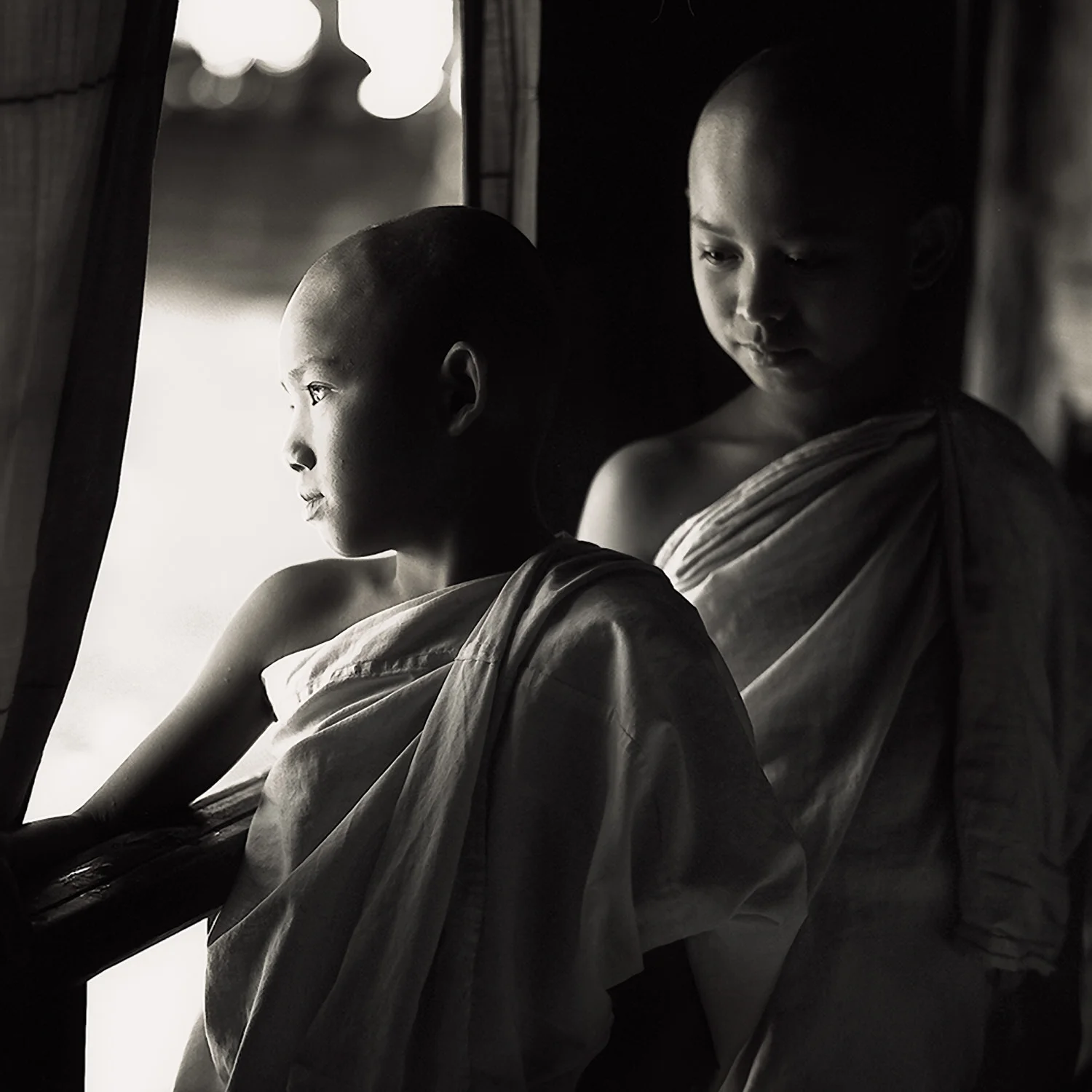 Two young novice monks , lit by side light , standing on a verandah in a monastry in Myanmar .