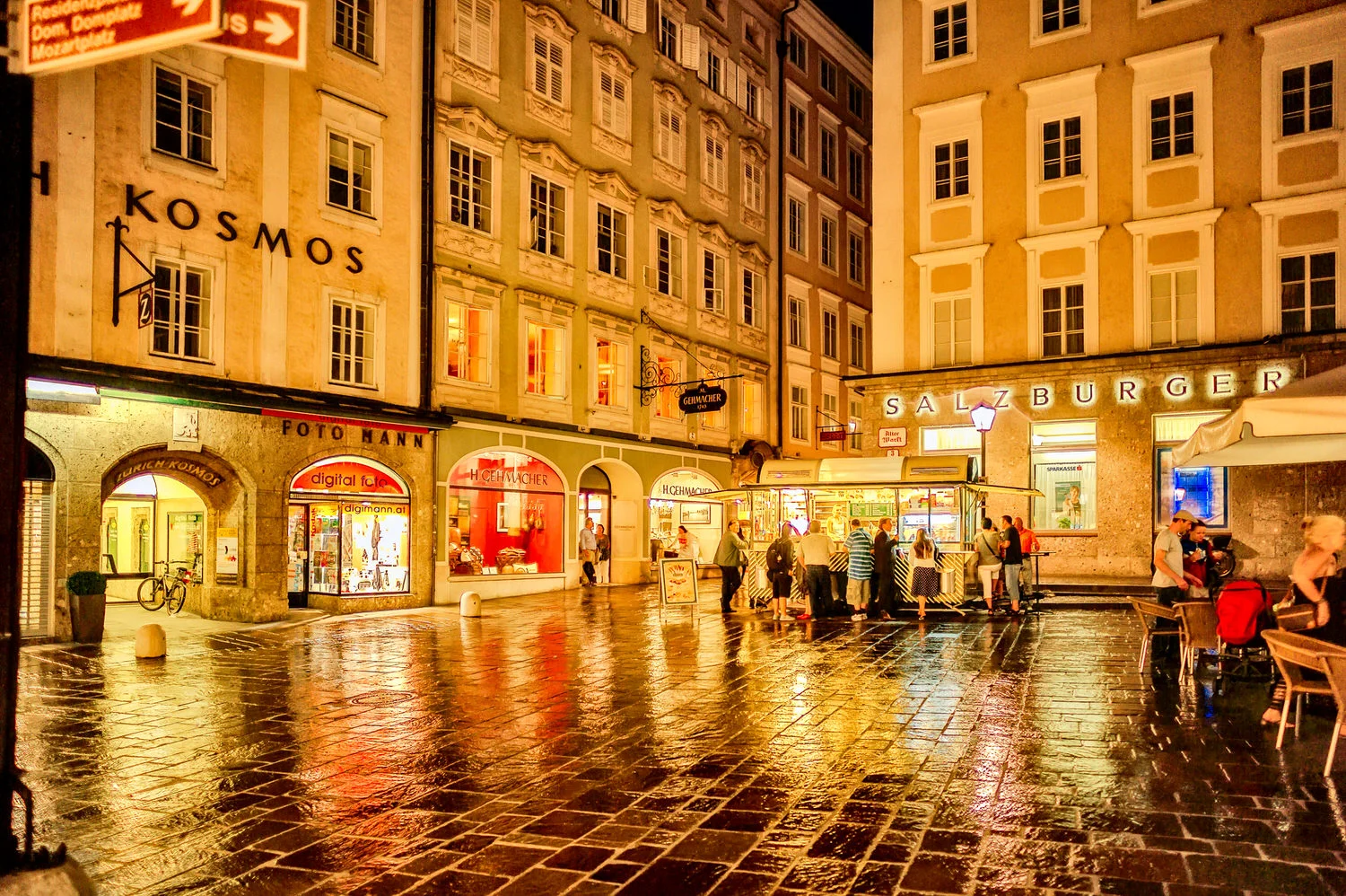 People enjoying a drink amidst a vividly lit street scene in the old town of Salzburg on a rainy summer's evening.