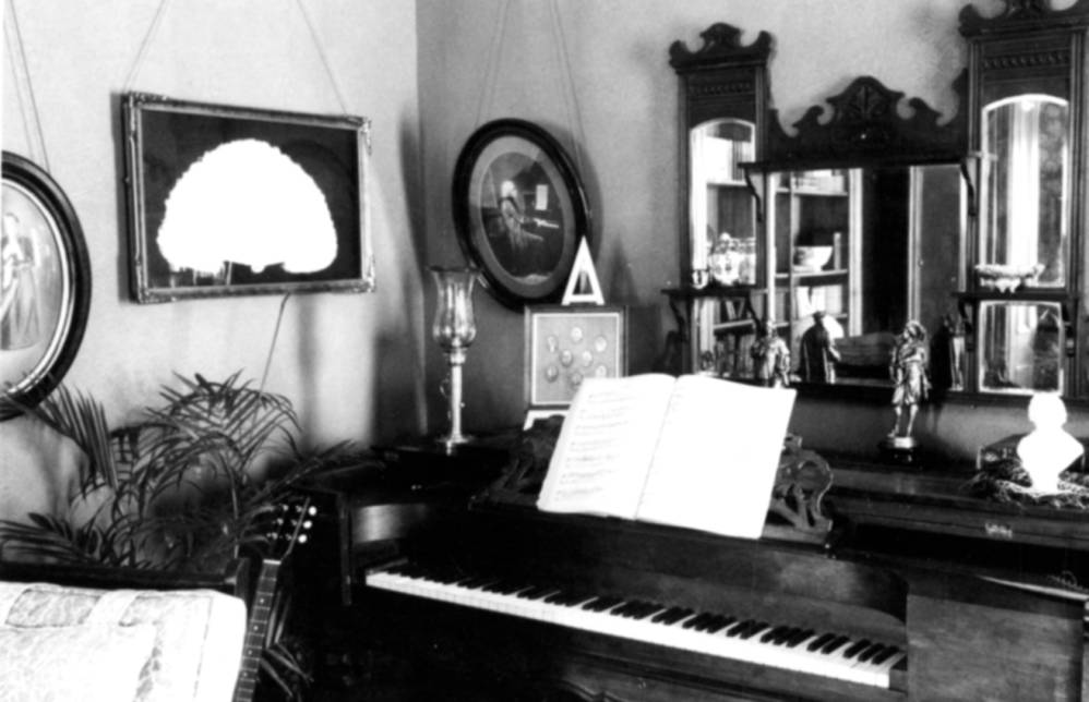  Circa 1980. This photo shows a close view of the piano against the wall in the corner of a room and the decorations on and around it. 