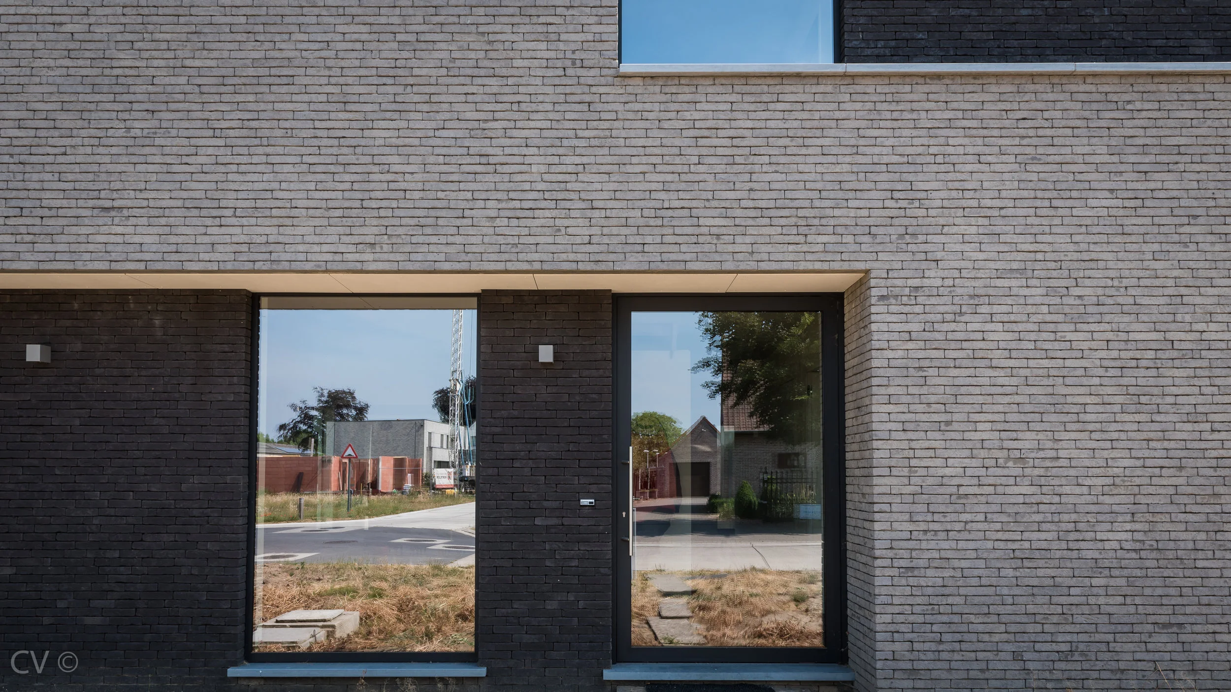 De bouwheer van deze nieuwe woning is een jong dynamisch koppel. Zij wensten een moderne ruime woning met specifiek aandacht voor oververhitting door de zuidoriëntatie. De carport moest mee geïntegreerd worden in het ontwerp. Vanaf het eerst voorontwerp werd geopteerd om carport en zonneluifel als 1 geheel te ontvouwen rondom een strak volume. Zowel binnen als in de luifel buiten werd een vide gecreëerd om interactie, zicht en licht toe te laten in de woning tussen buiten/binnen en beneden/boven. Op het gelijkvloers wordt gewerkt met een open plan voor keuken, eetruimte en leefruimte. De trap naar de verdieping komt ook uit in de leefruimte. Er werd ook ruimte voorzien voor een bureel voor de bewoners. Op de verdieping zijn 3 ruime slaapkamers uitgewerkt, een dressing, een archiefruimte en een badkamer. Het ontwerp resulteerde uiteindelijk in een BEN-woning E-peil : E16 K-peil : K38