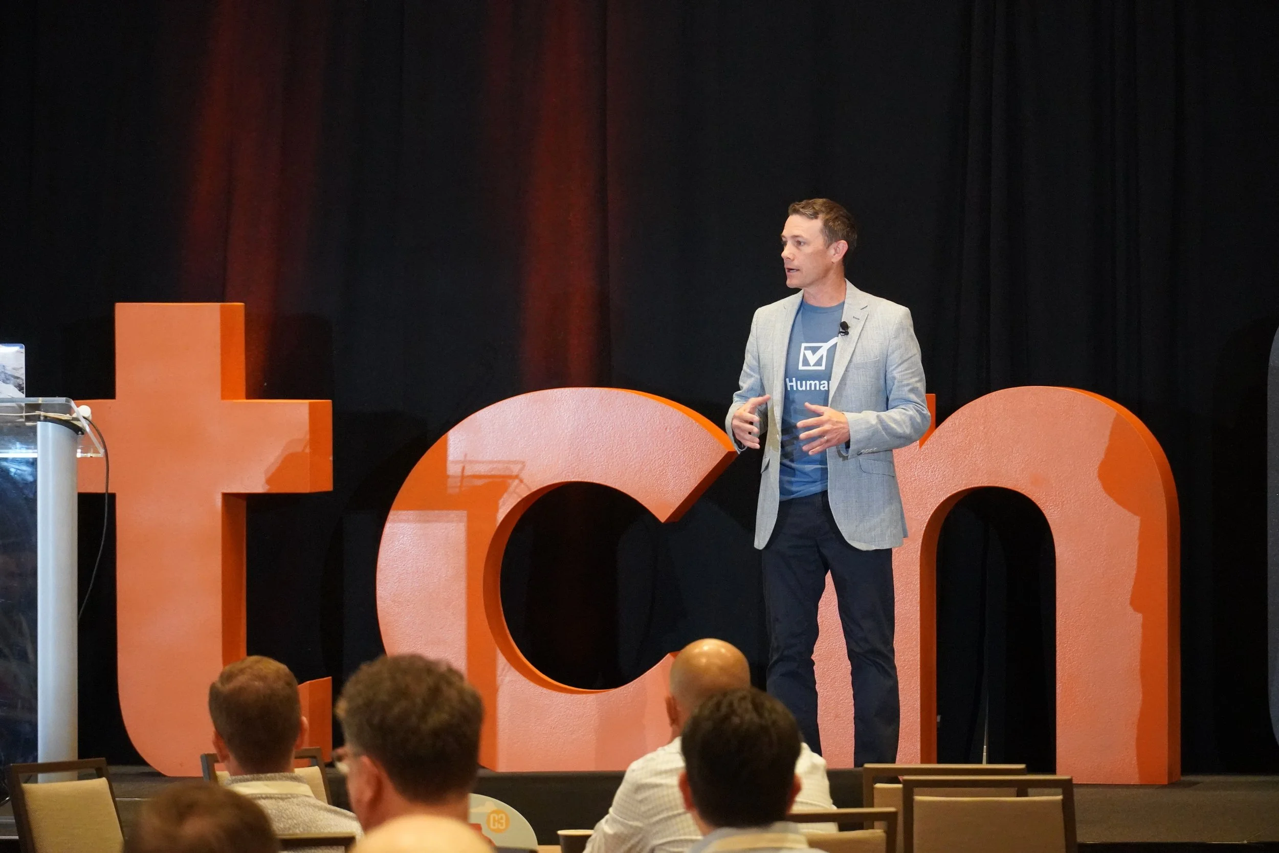 Keynote speaker Jeff Toister is presenting from the stage. He is wearing a blazer with a t-shirt underneath that reads "human." The letters T,C,N are on the stage in the background.