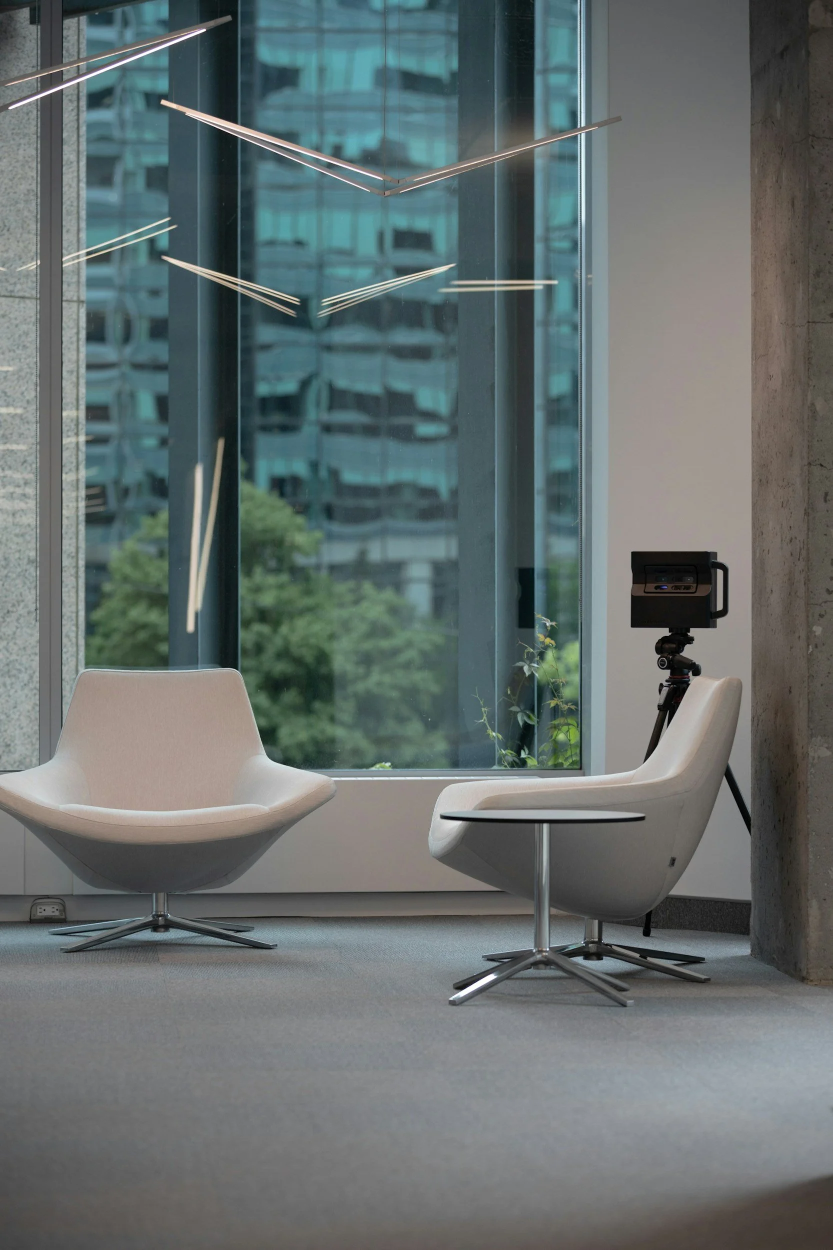 Modern office interior with two white lounge chairs near large window, a small black camera or projector on a tripod, and reflections of city buildings outside.