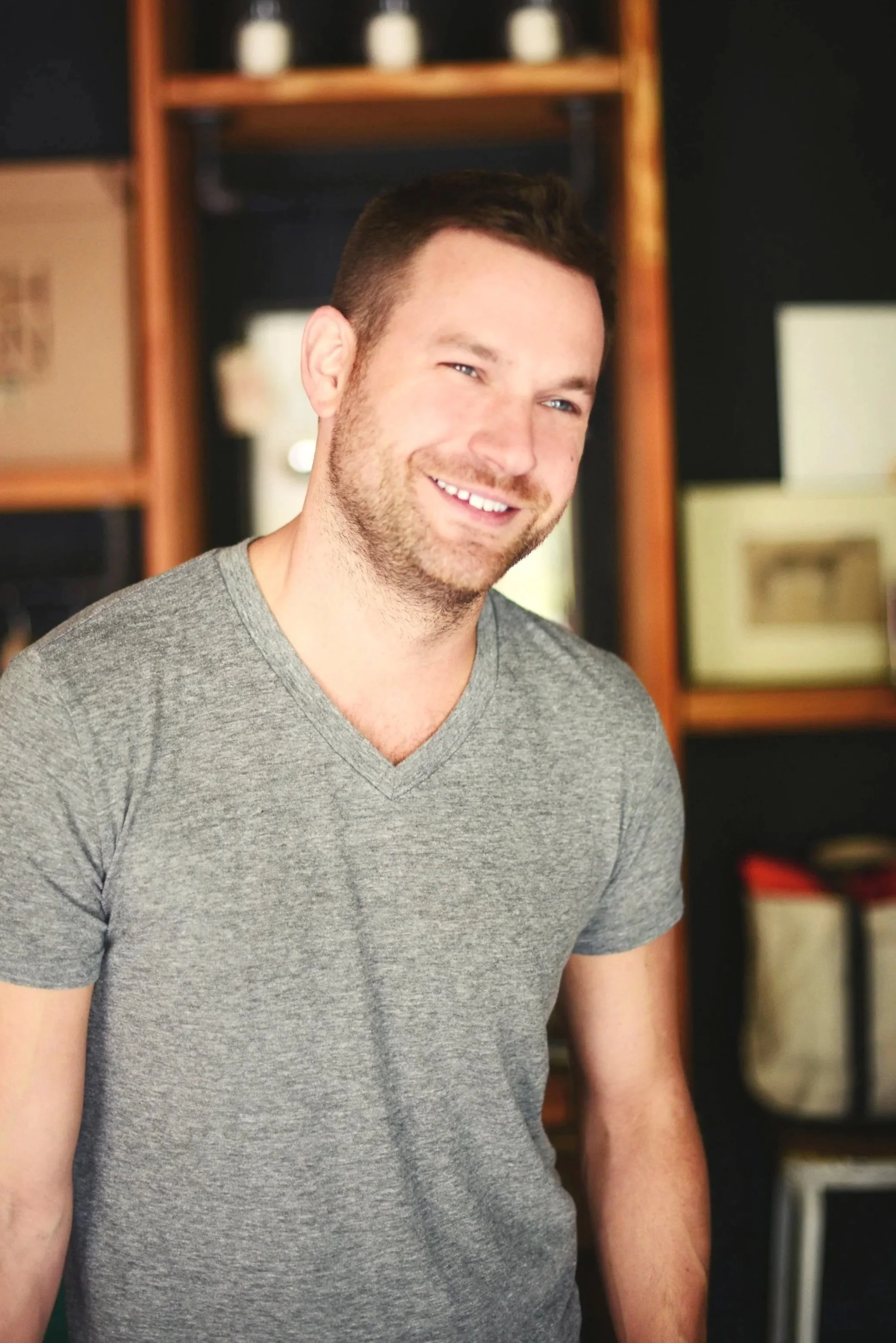 A young man with short dark hair, a light beard, and a gray T-shirt, smiling in a room with wooden shelves and framed pictures in the background.