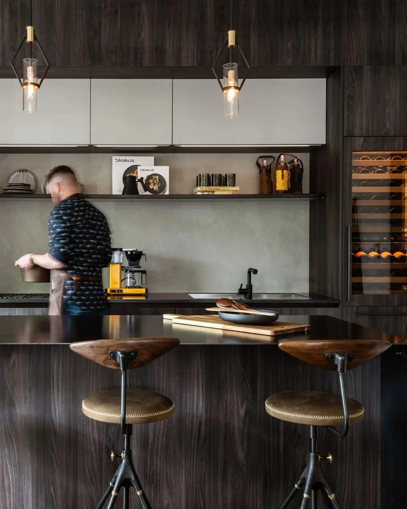 Modern kitchen with dark wooden finish, white upper cabinets, and open shelves with kitchenware. A man is preparing coffee, surrounded by a coffee machine and utensils. There are two wooden barstools at a dark wood counter.