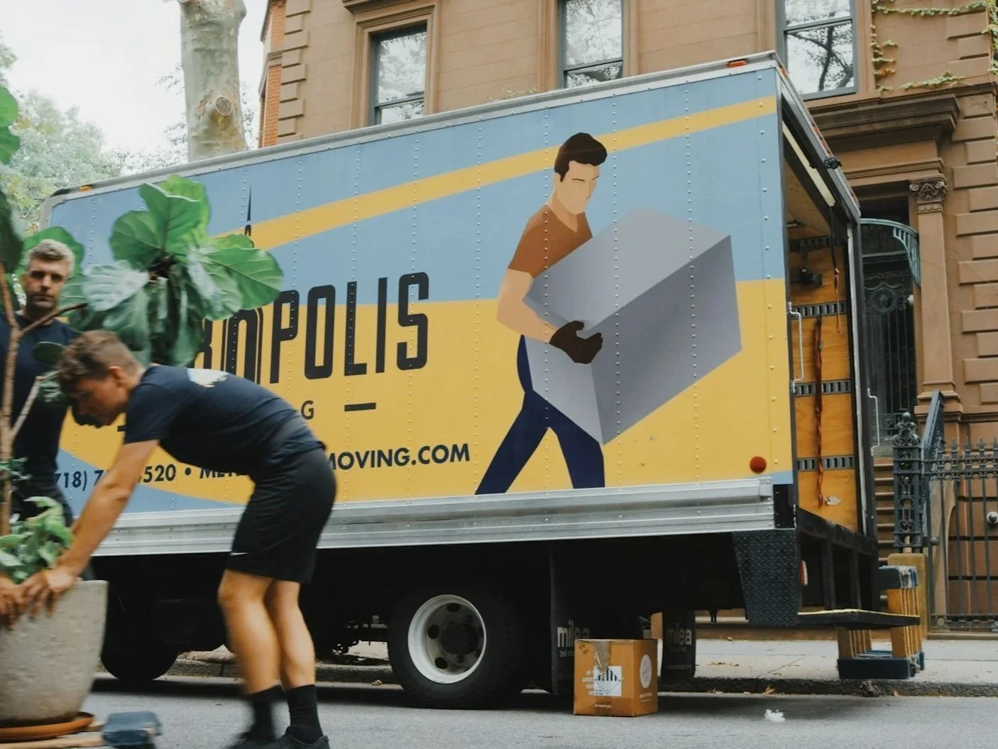 People moving plants into a moving truck with city buildings in the background. The truck has an advertisement showing a person carrying a box and the words 'POLIS' and 'MOVING.COM'.