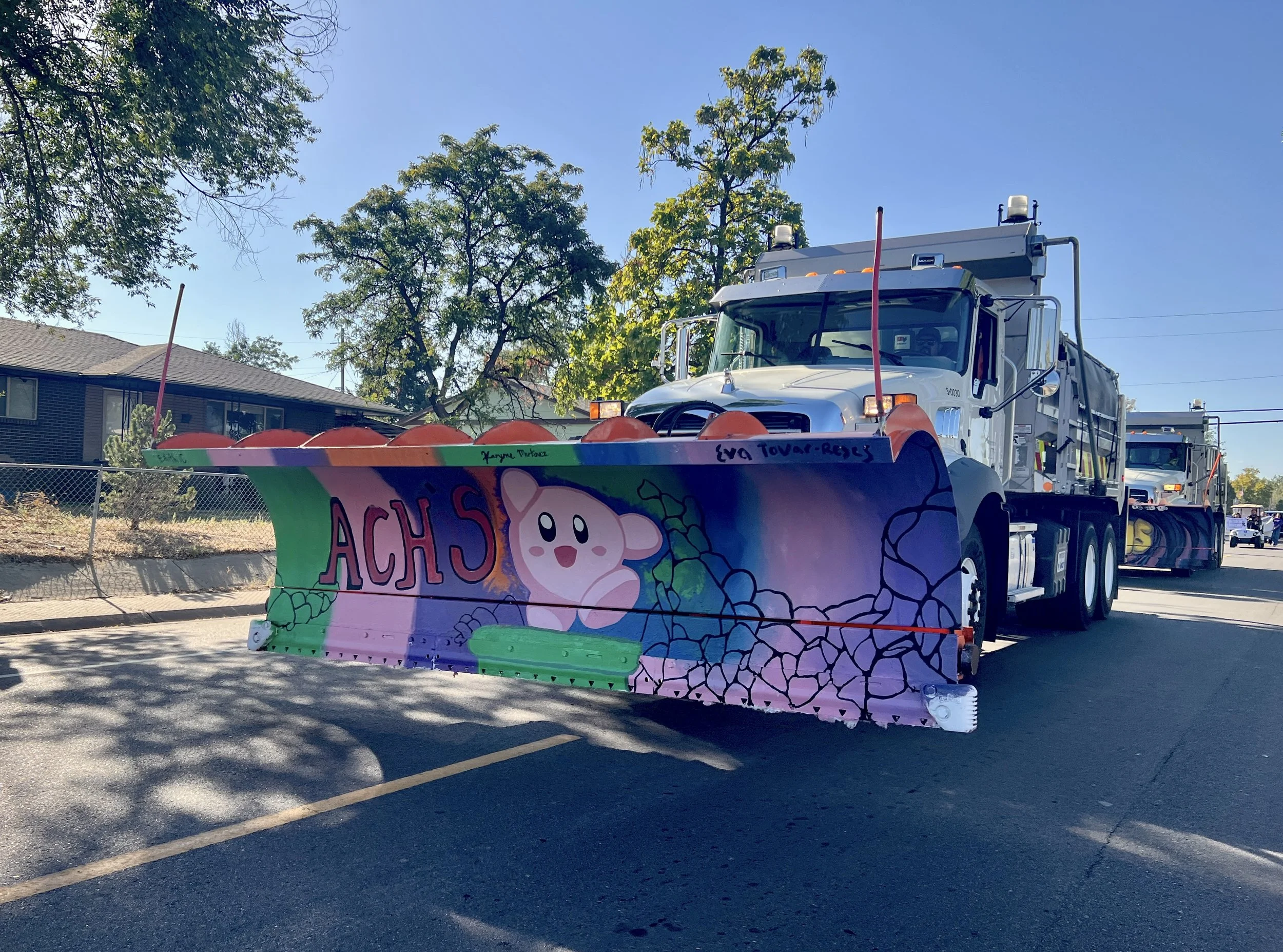 A parade float with colorful artistic designs, including a cute cartoon character and the text 'ACH5', being pulled by a large white truck on a sunny street with trees and residential buildings in the background.
