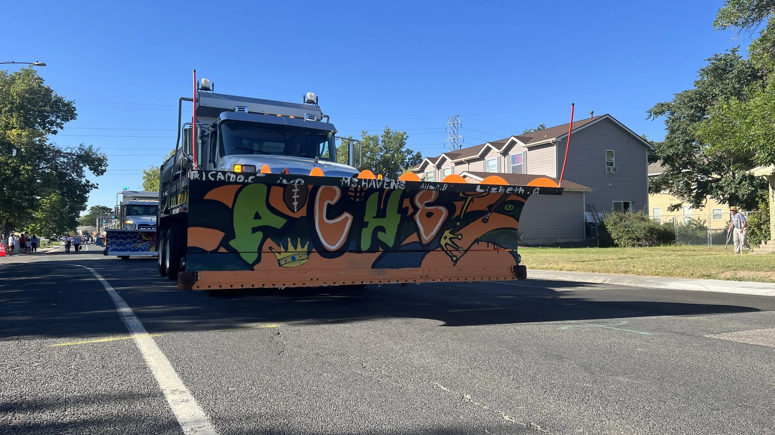 A street scene with a black street sweeper decorated with colorful graffiti art on its front, sweeping the road for a parade or event, with houses, trees, and people in the background.