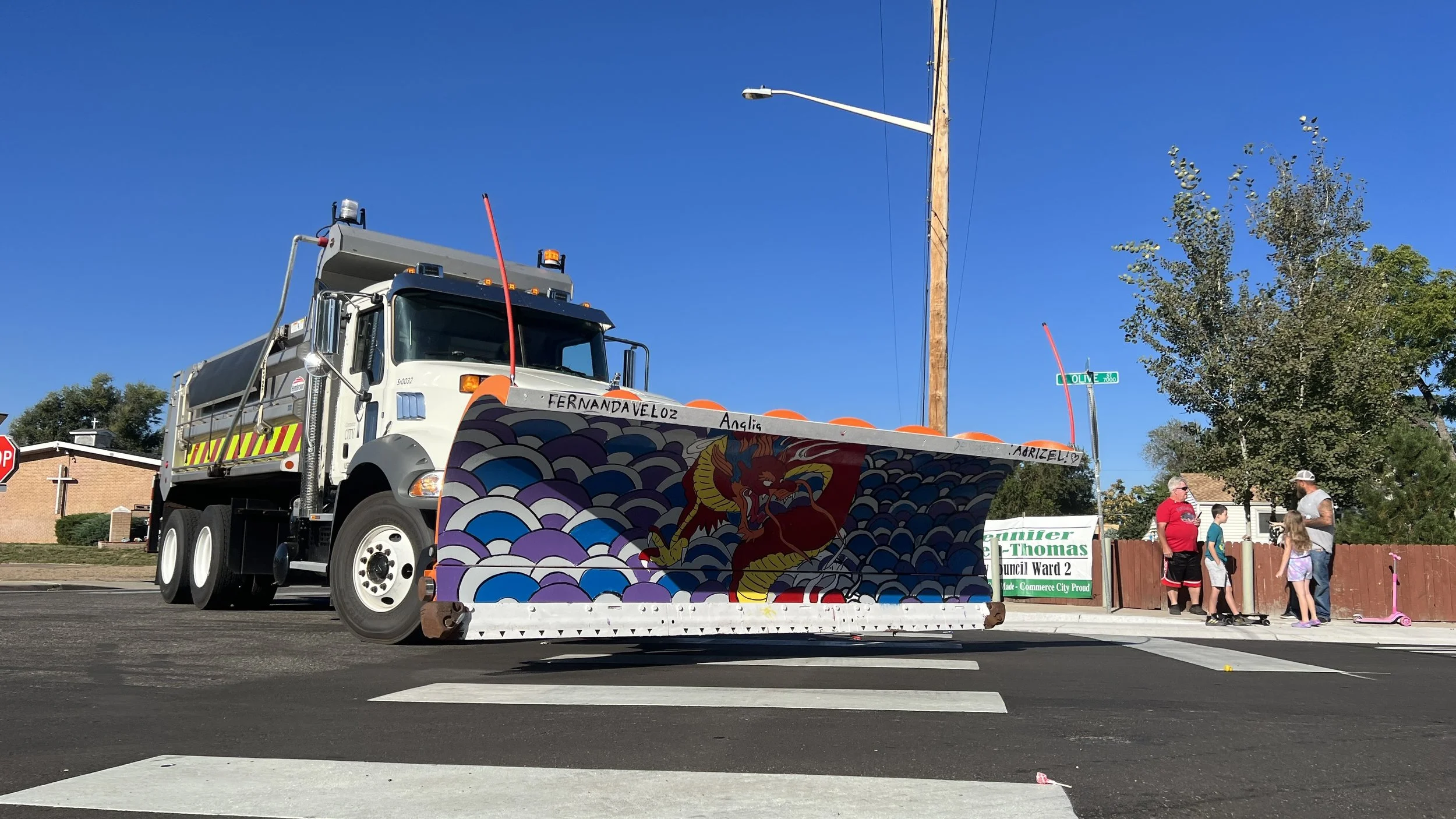 A street scene with a large water truck and a decorated float featuring a red and yellow dragon and blue waves, with people standing and talking near a sign that reads 'Walker Thomas Council Ward 2' on a sunny day.