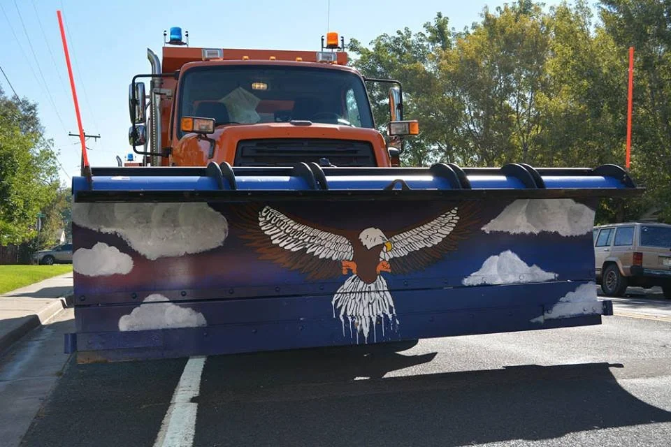 An orange snowplow with a painted front blade featuring an eagle flying among clouds, parked on a street with trees and other vehicles in the background.