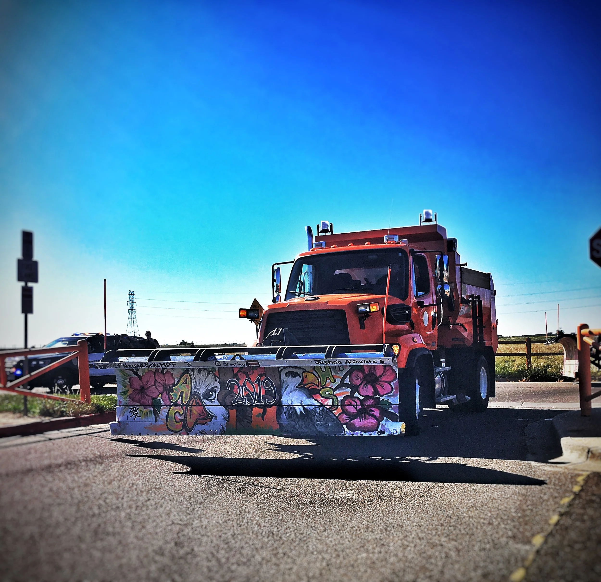 A red street sweeping truck with graffiti artwork on its front blade, parked on a street under a clear blue sky.