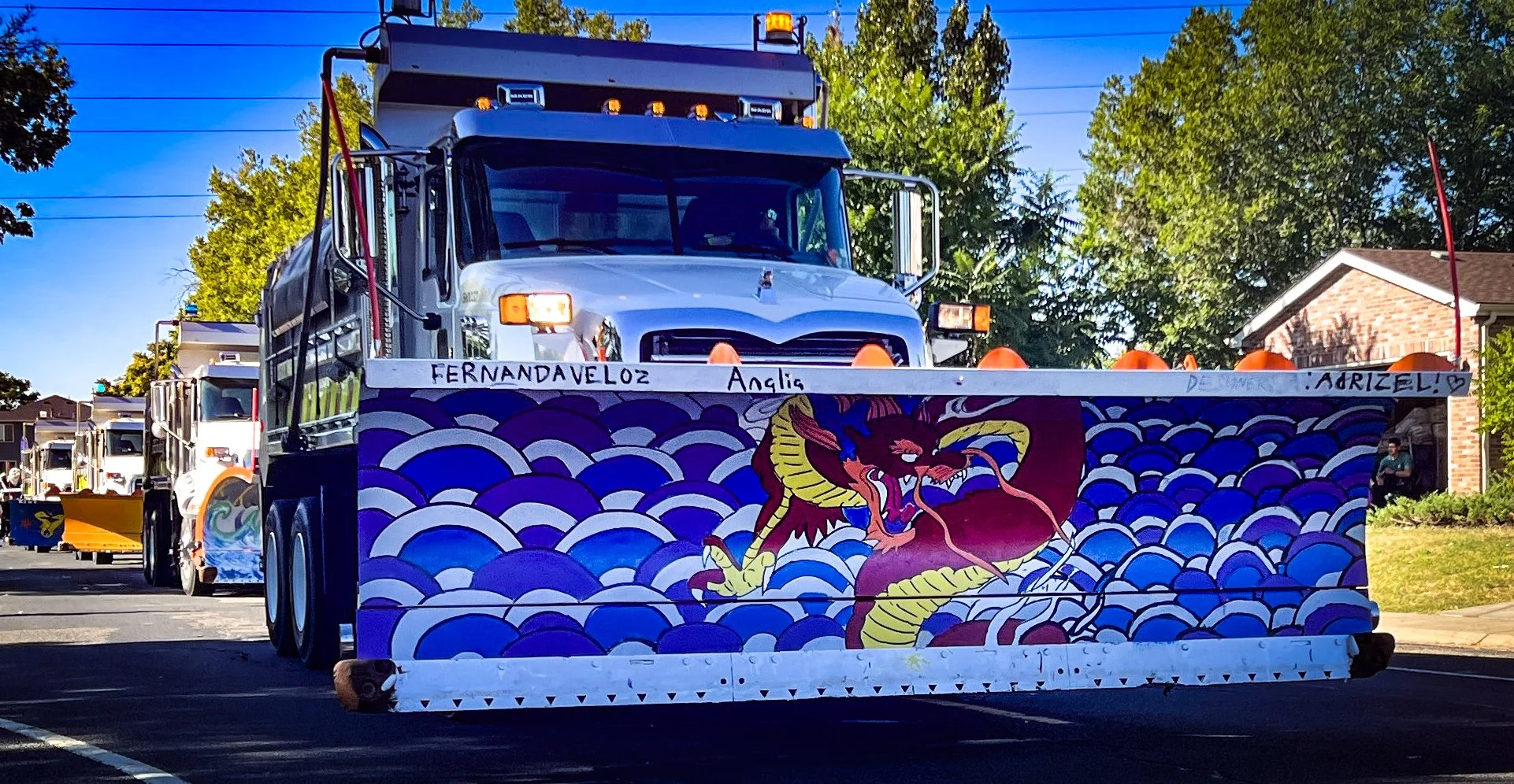 A parade float decorated with colorful artwork of a dragon against a wave pattern, with handwritten names and messages, being pulled through a neighborhood street lined with trees and houses.