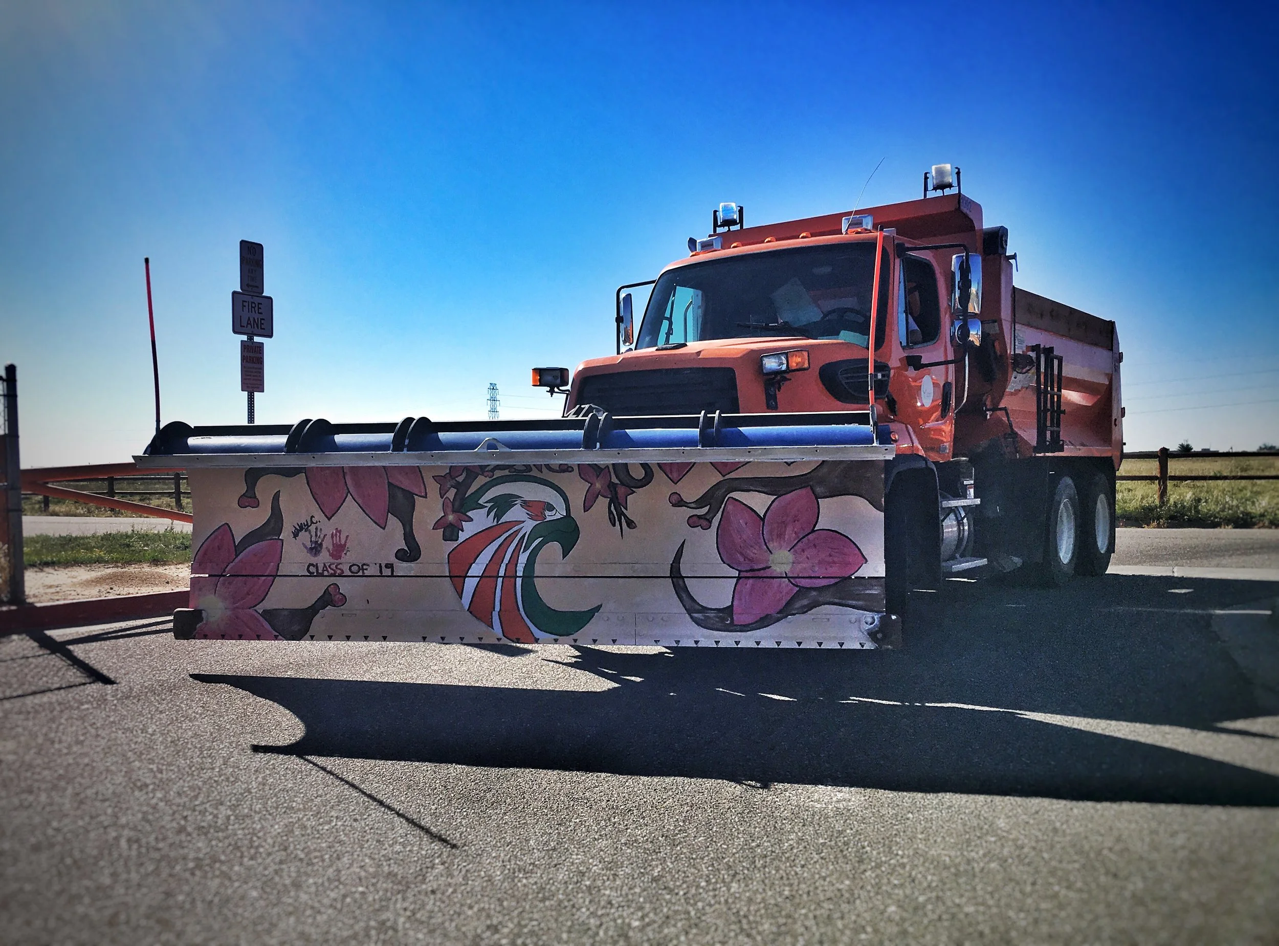A red street plow truck with pink floral and eagle decorations on the front, parked on the side of the road next to a field and clear blue sky.